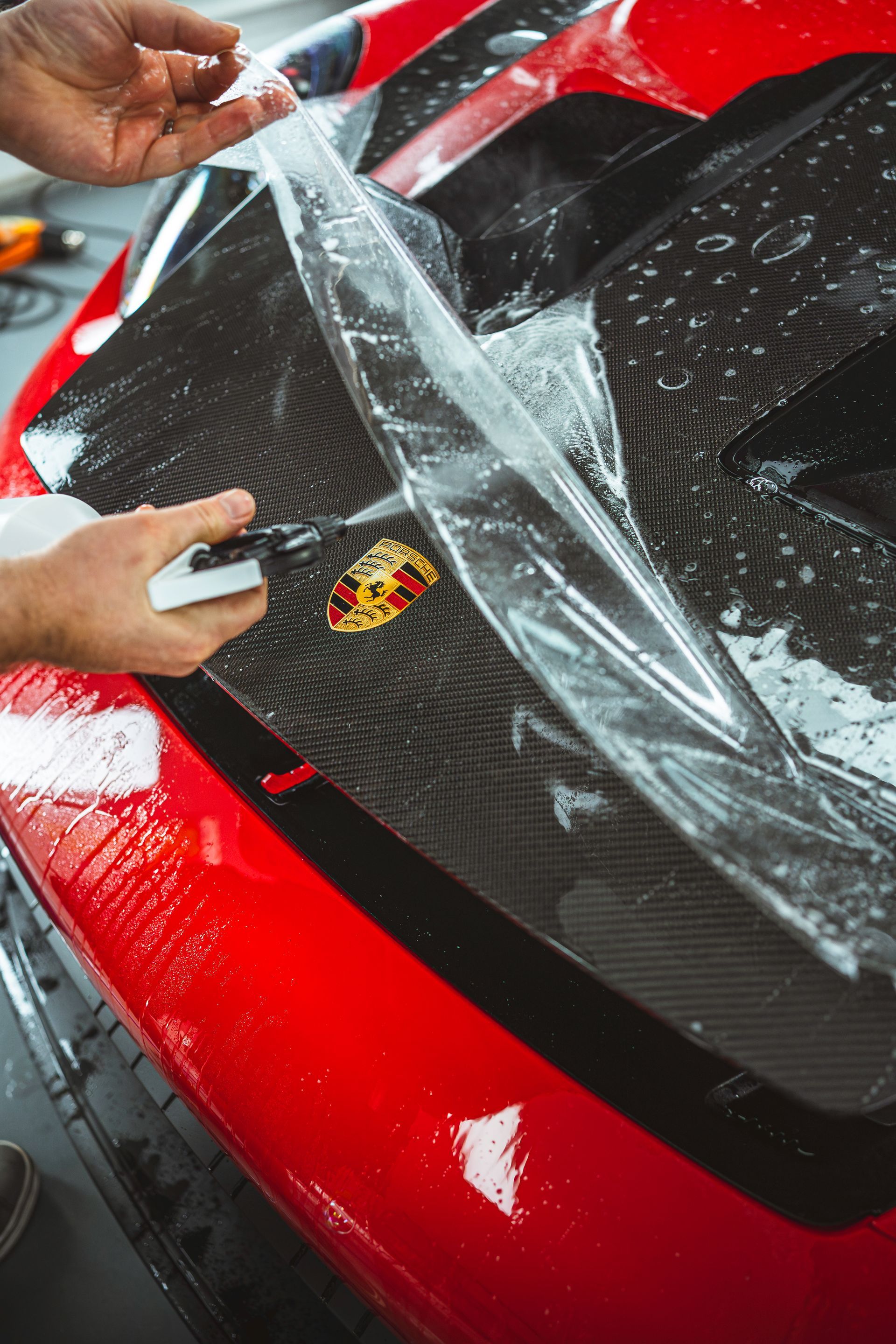 Hands peeling protective film from a red sports car hood with a Porsche emblem