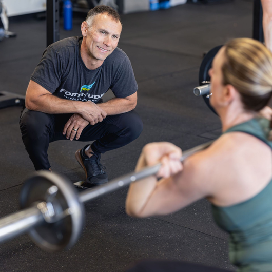 Coach crouching beside a person lifting a barbell in a gym, offering guidance