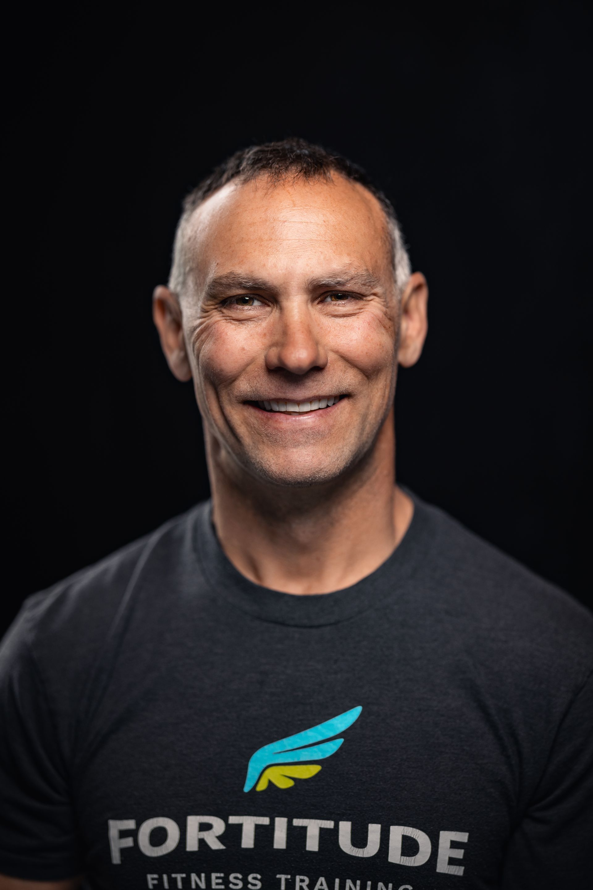 Smiling man in a dark T-shirt with “Fortitude” logo against a black background