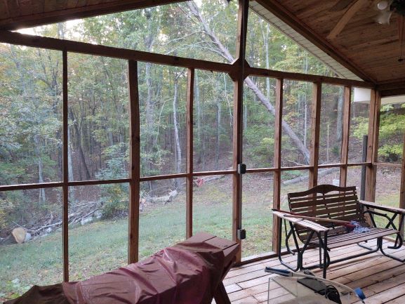A screened in porch with a bench and chairs and a view of the woods.