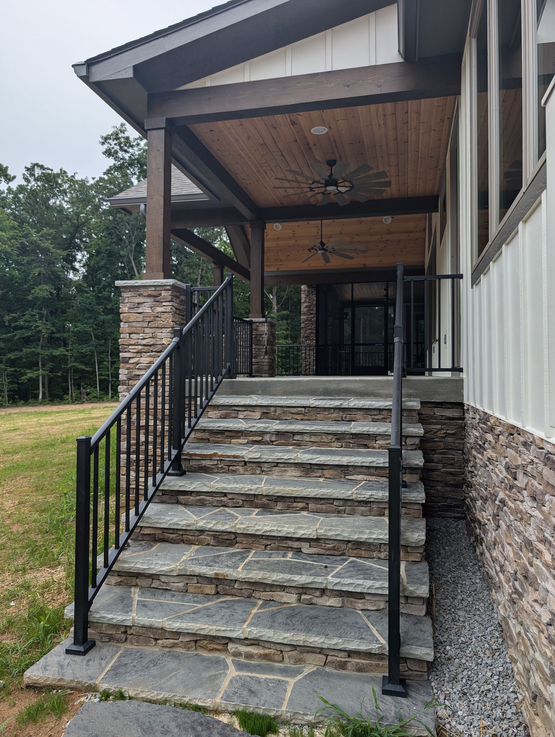 Stone staircase leading to a covered porch with black railing and stone columns on a house exterior