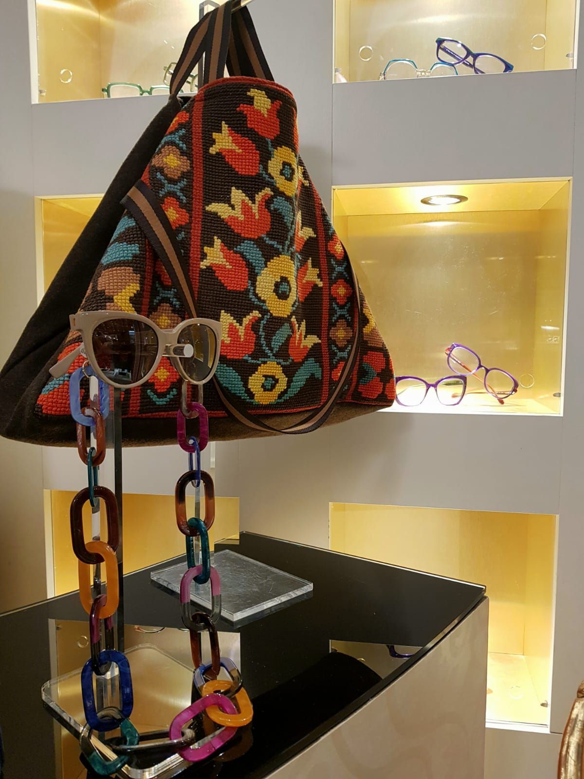 Borsa a mano colorata ricamata esposta su uno scaffale con occhiali e tracolla a catena multicolore