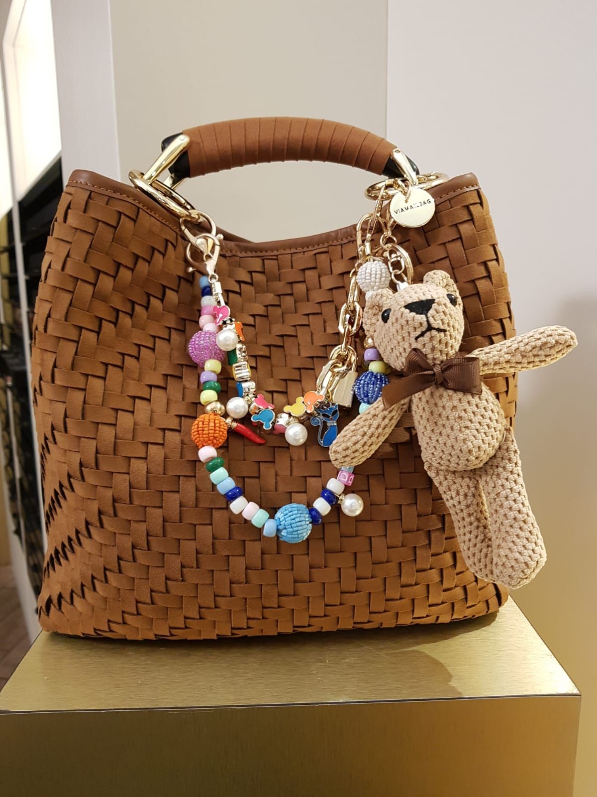 Borsa a mano marrone intrecciata con collana di perline colorate e piccolo ciondolo a forma di orsetto appeso sul davanti.