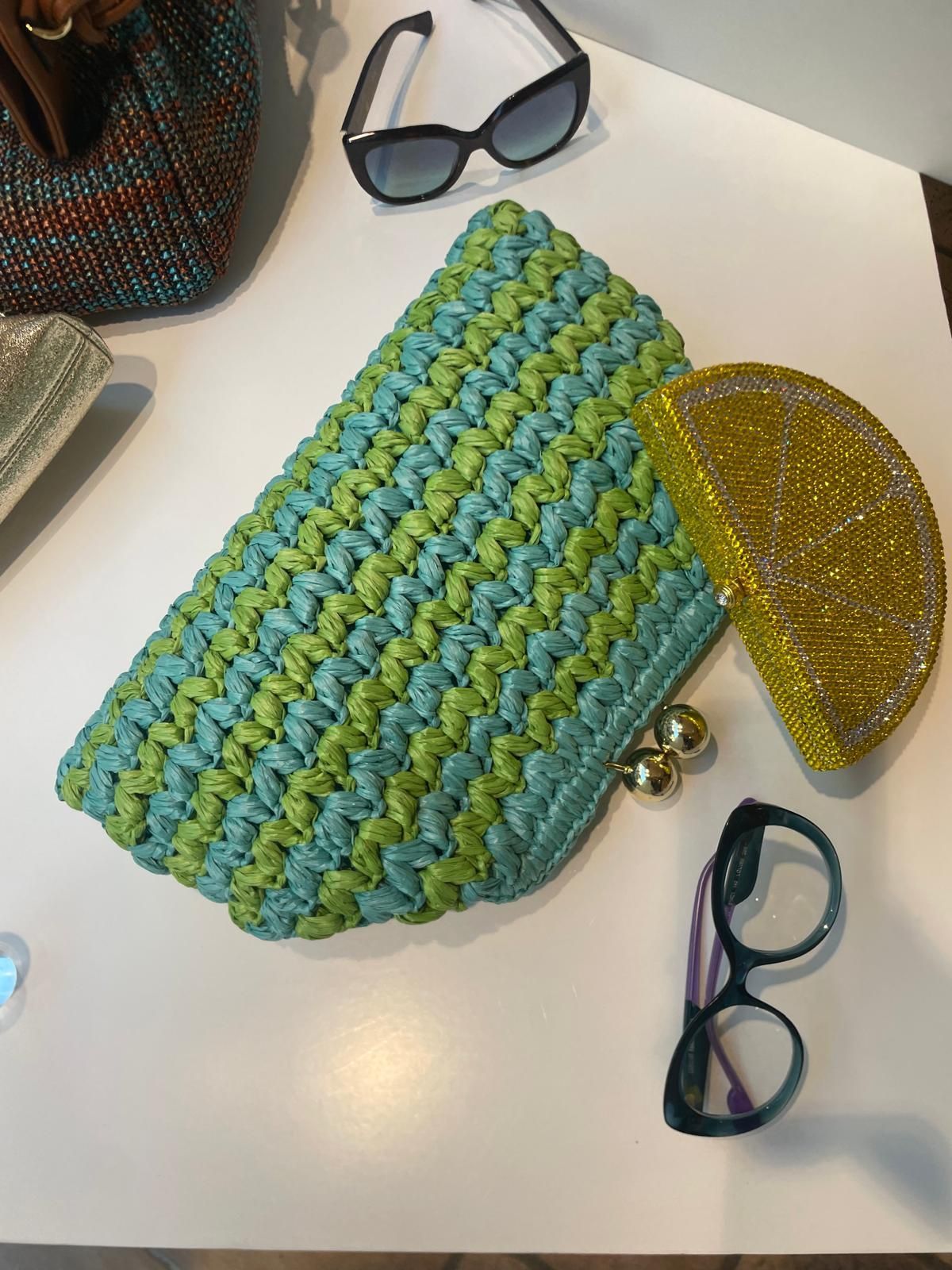Pochette intrecciata blu-verde con borsetta a mezzaluna gialla decorata con perline e occhiali da sole su un tavolo bianco.