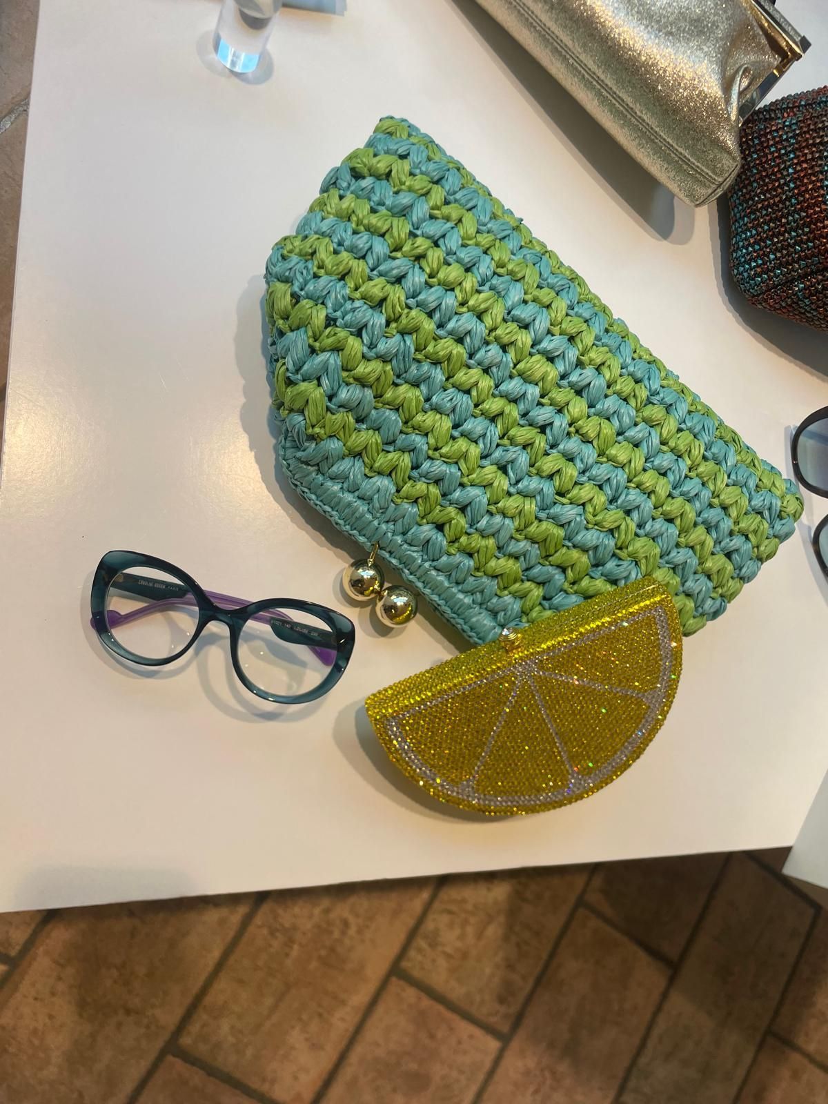 Pochette intrecciata blu-verde, pochette a mezzaluna gialla scintillante e occhiali rotondi su una superficie bianca.