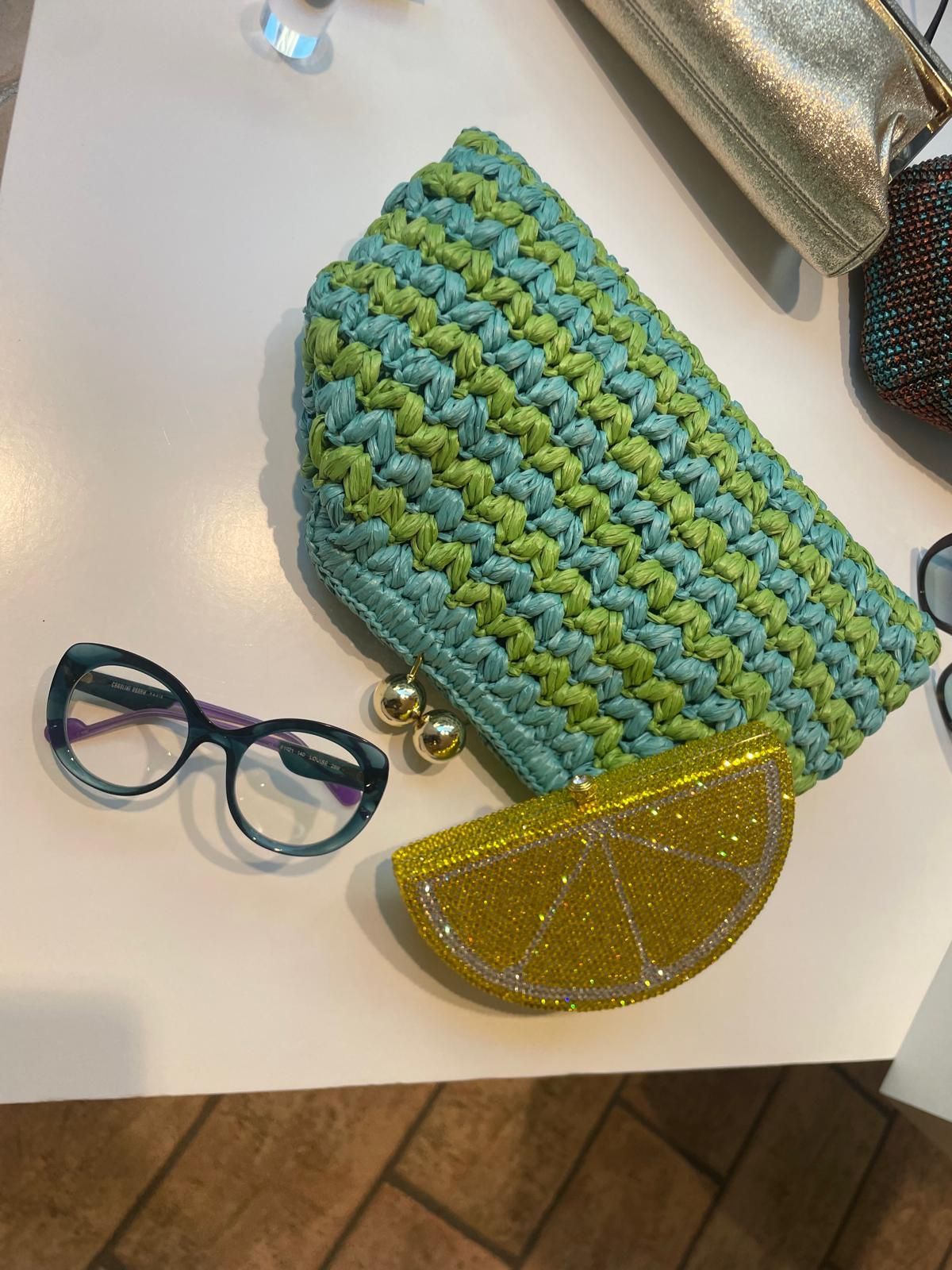 Una pochette intrecciata blu-verde, un portamonete giallo e bicchieri rotondi su un tavolo chiaro.