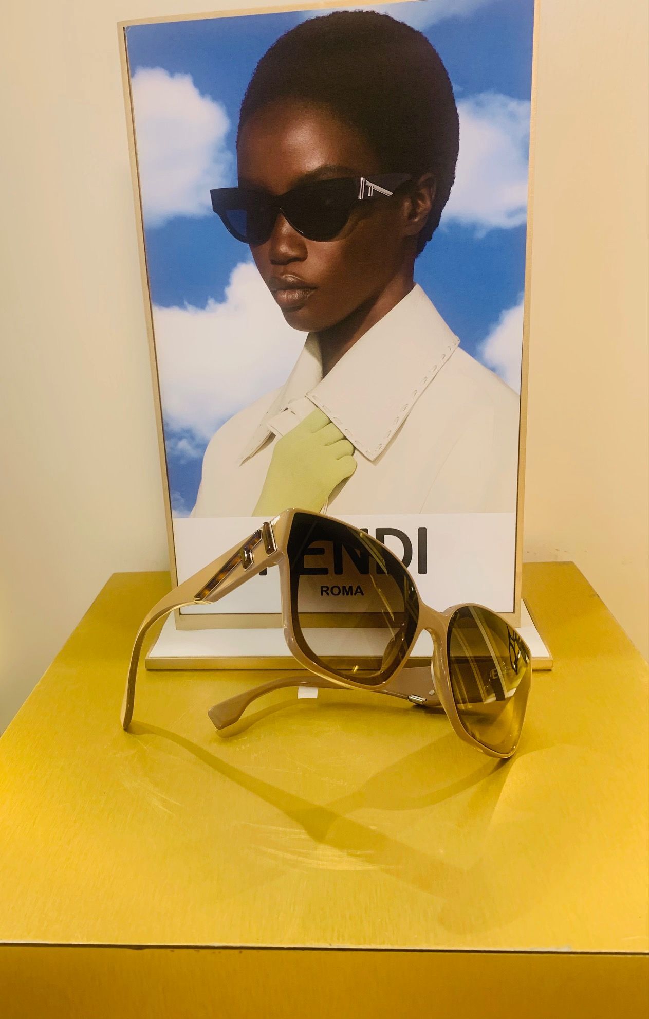 Occhiali da sole dorati su un espositore giallo con un poster di moda sullo sfondo.