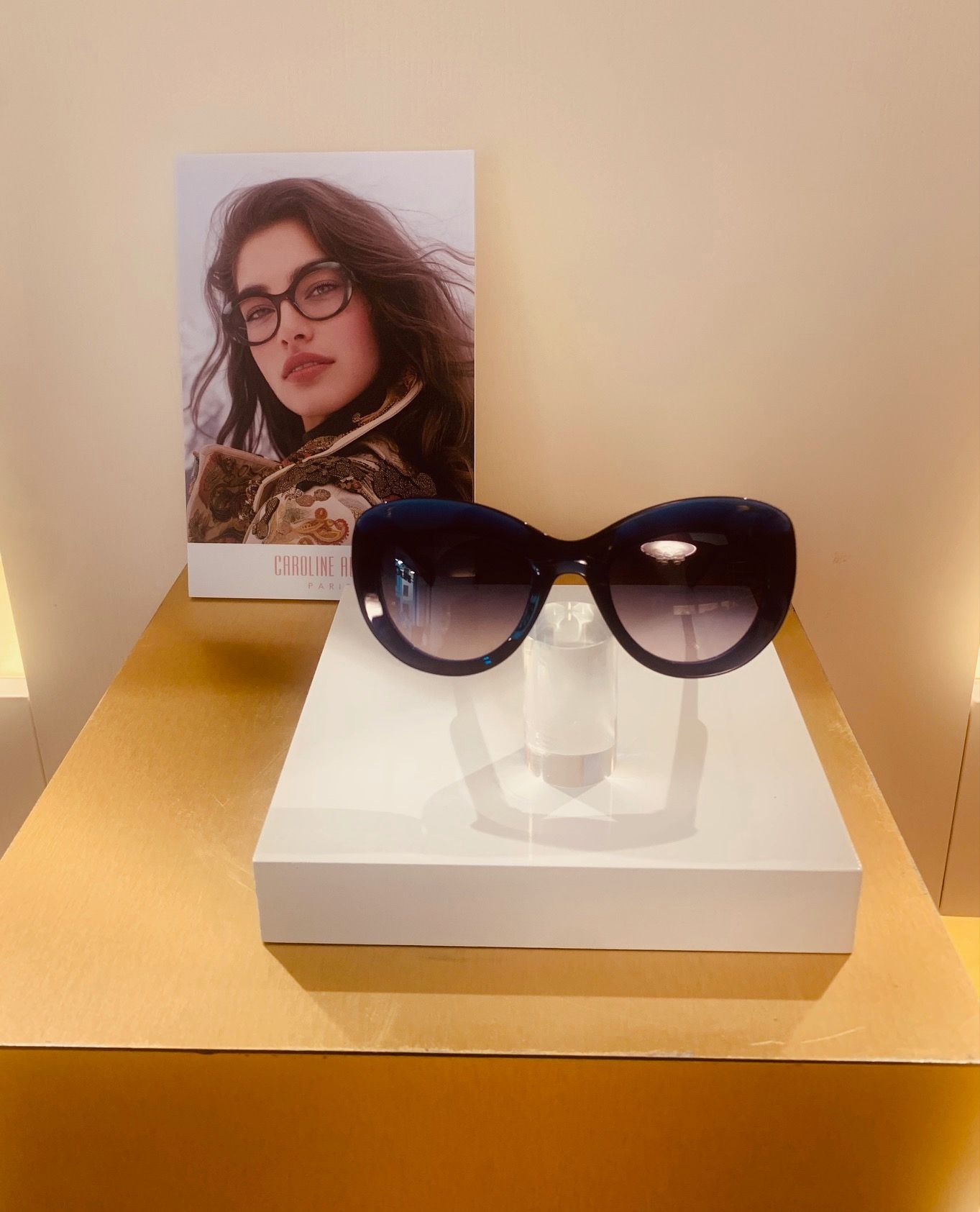 Occhiali da sole su un espositore bianco accanto a un poster di moda in una vetrina beige.