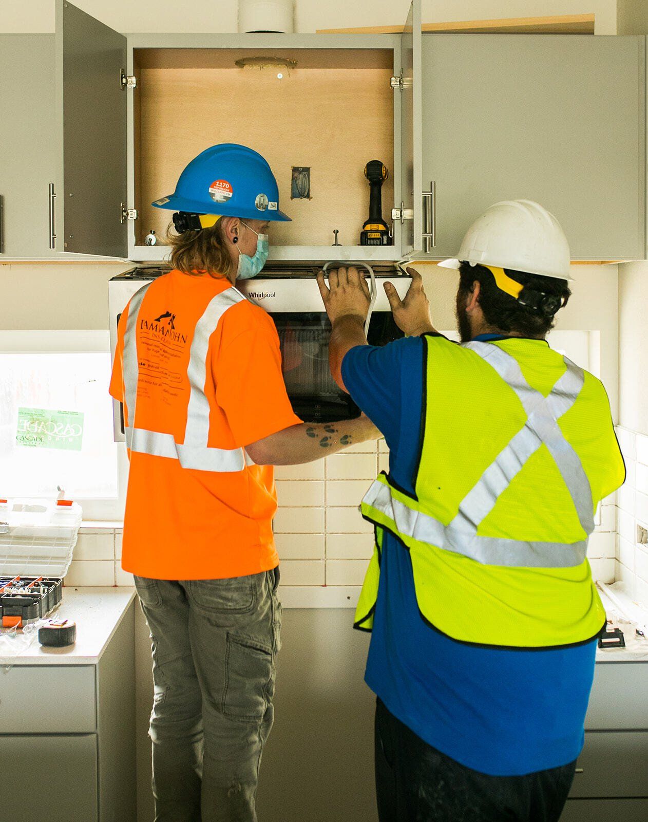 Two men wearing hard hats and safety vests are working in a kitchen