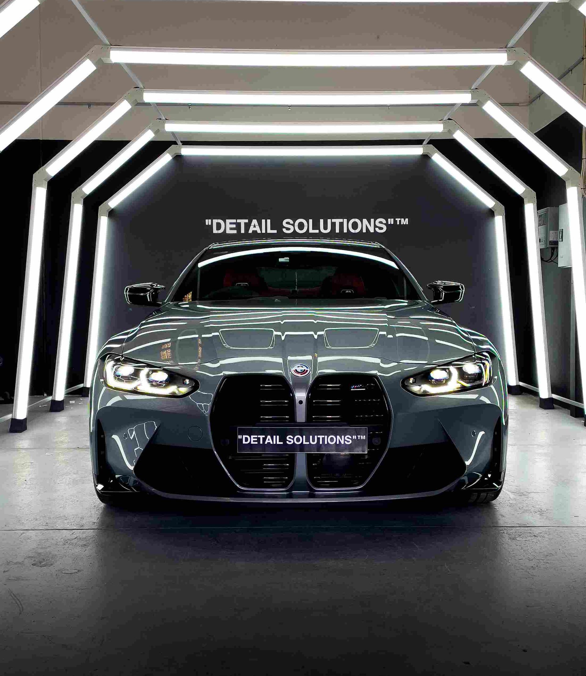 Gray BMW M4 coupe parked inside a detailing shop with bright lights, “Detail Solutions” logo visible.