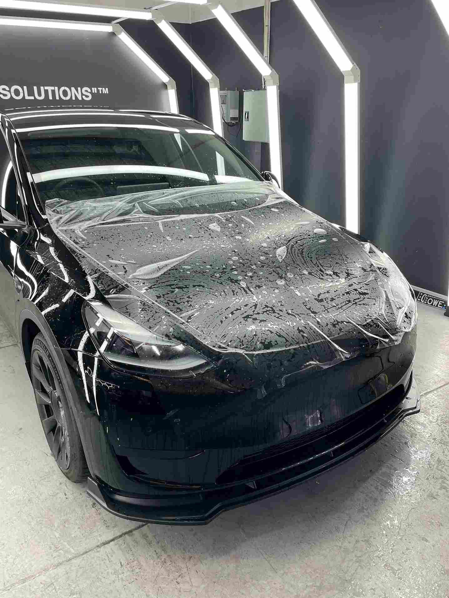 Black car with clear film on hood, in a well-lit shop.