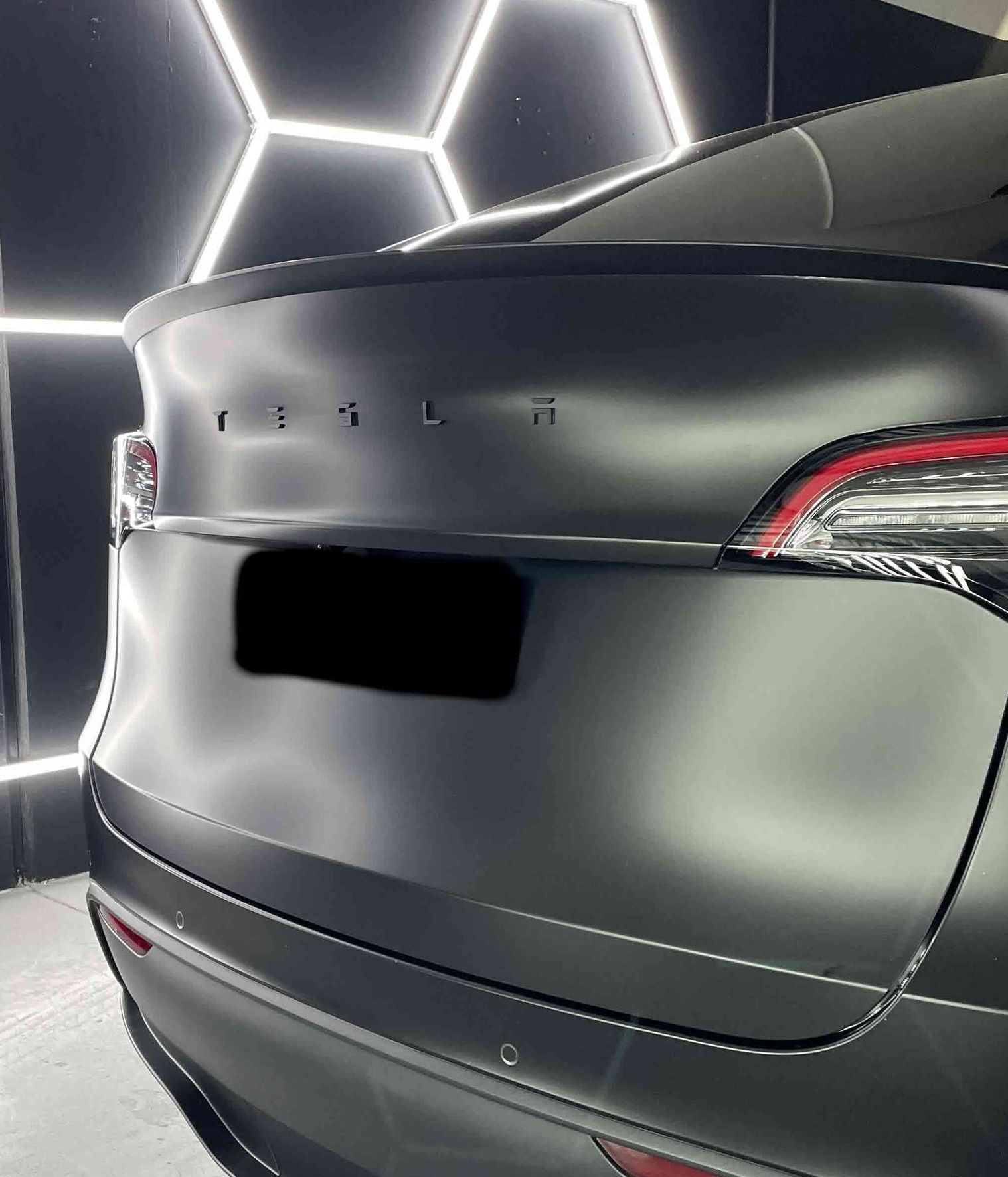 Matte black Tesla Model Y rear with lights on in a studio lit by hexagonal lights.