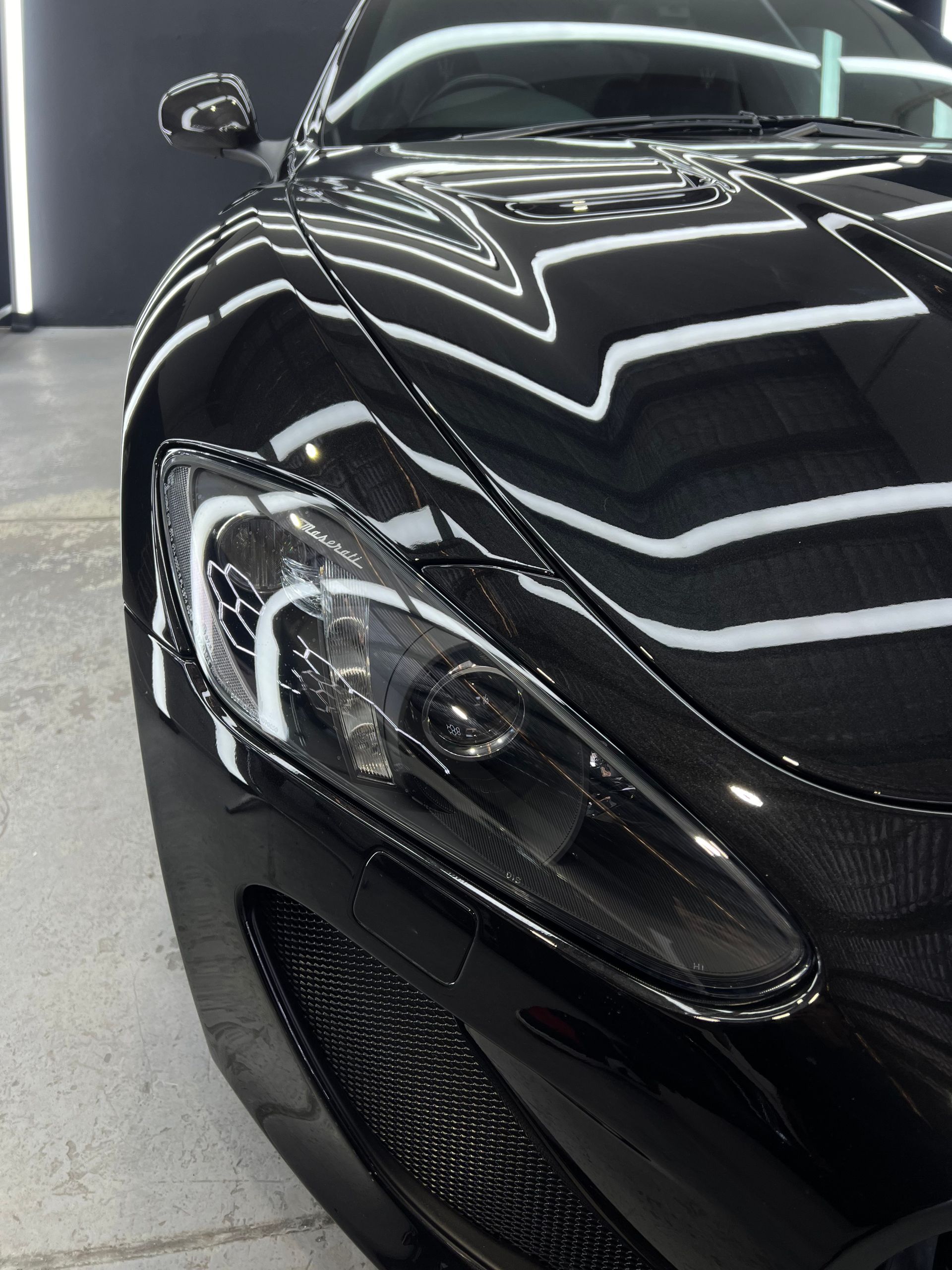 Black sports car's front with a reflective surface, headlight, and grille visible; indoors, likely at a detailing shop.