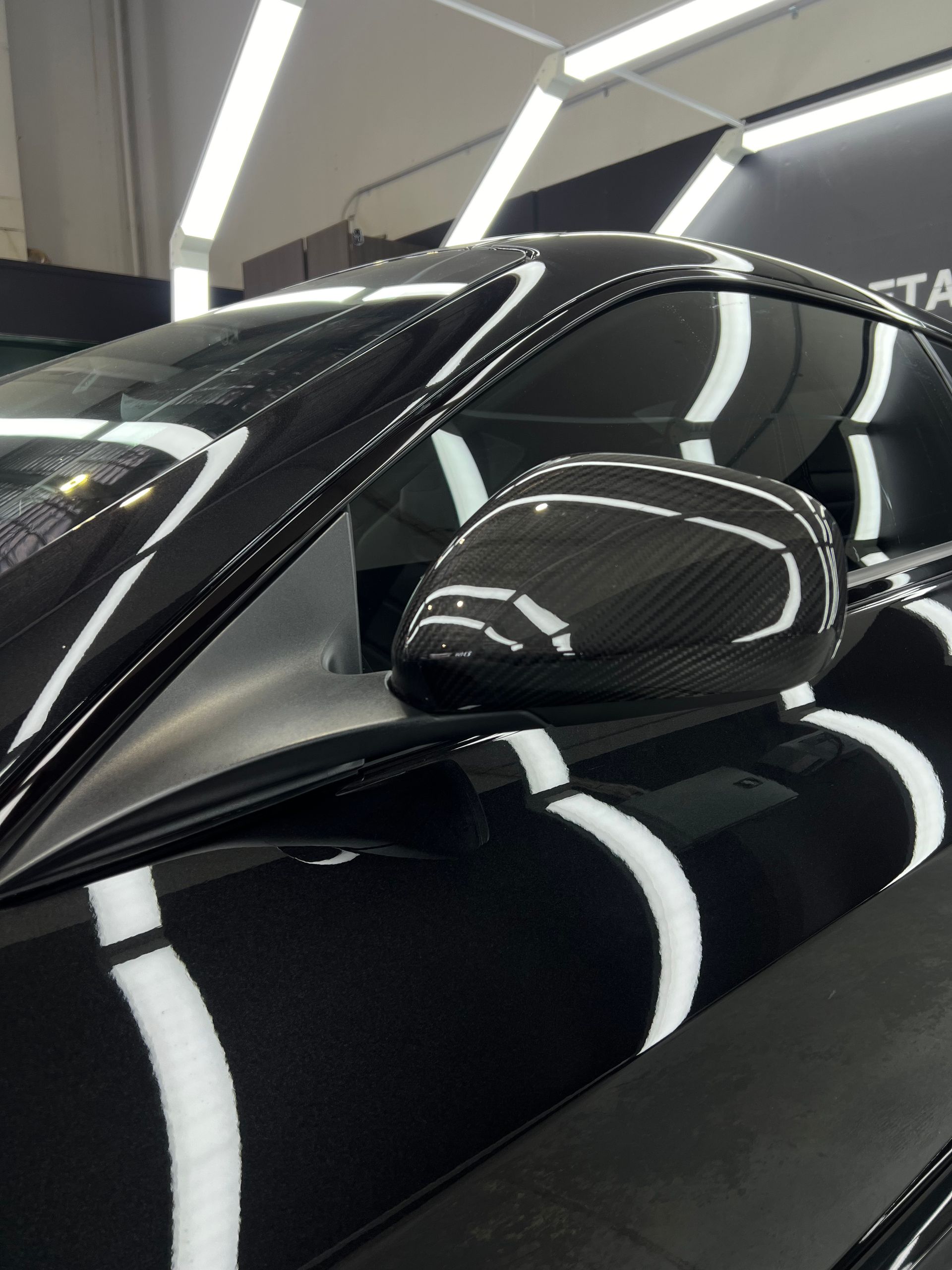 Black sports car with carbon fiber side mirror, under bright overhead lights.