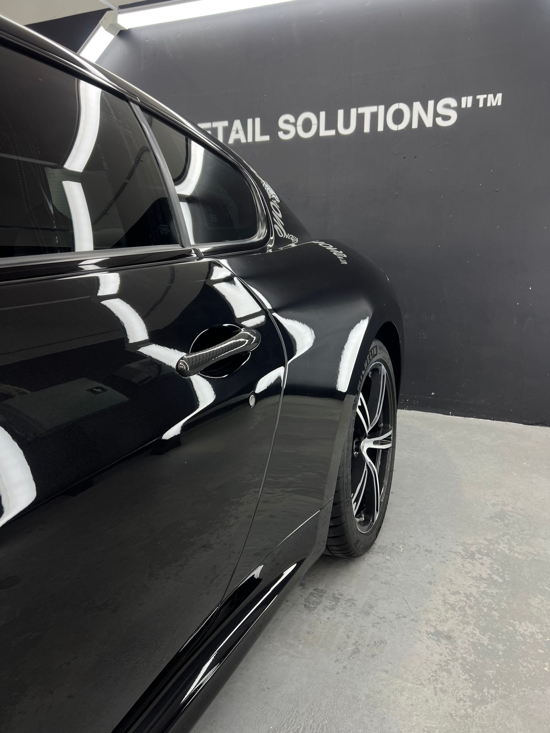 Black car, side view, reflecting lights, parked in a studio with “DETAIL SOLUTIONS” on the wall.
