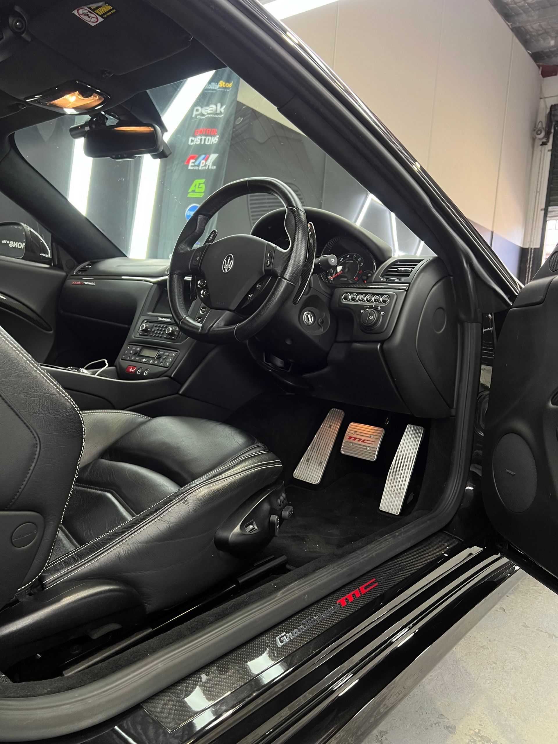 Black Maserati interior with black leather seats, steering wheel, and silver pedals.