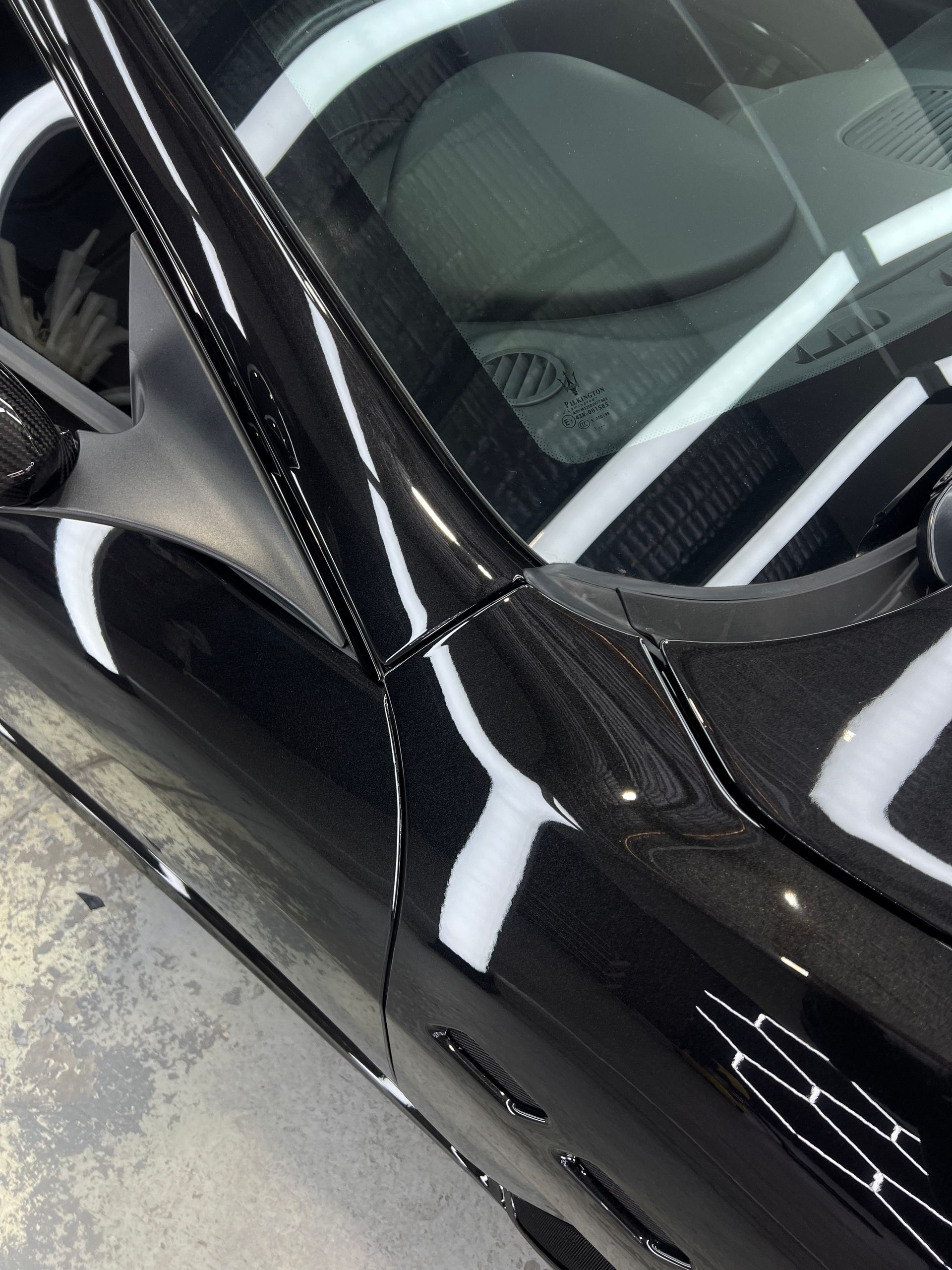 Close-up of a glossy black car's carbon fiber frame and windshield, reflecting white light.
