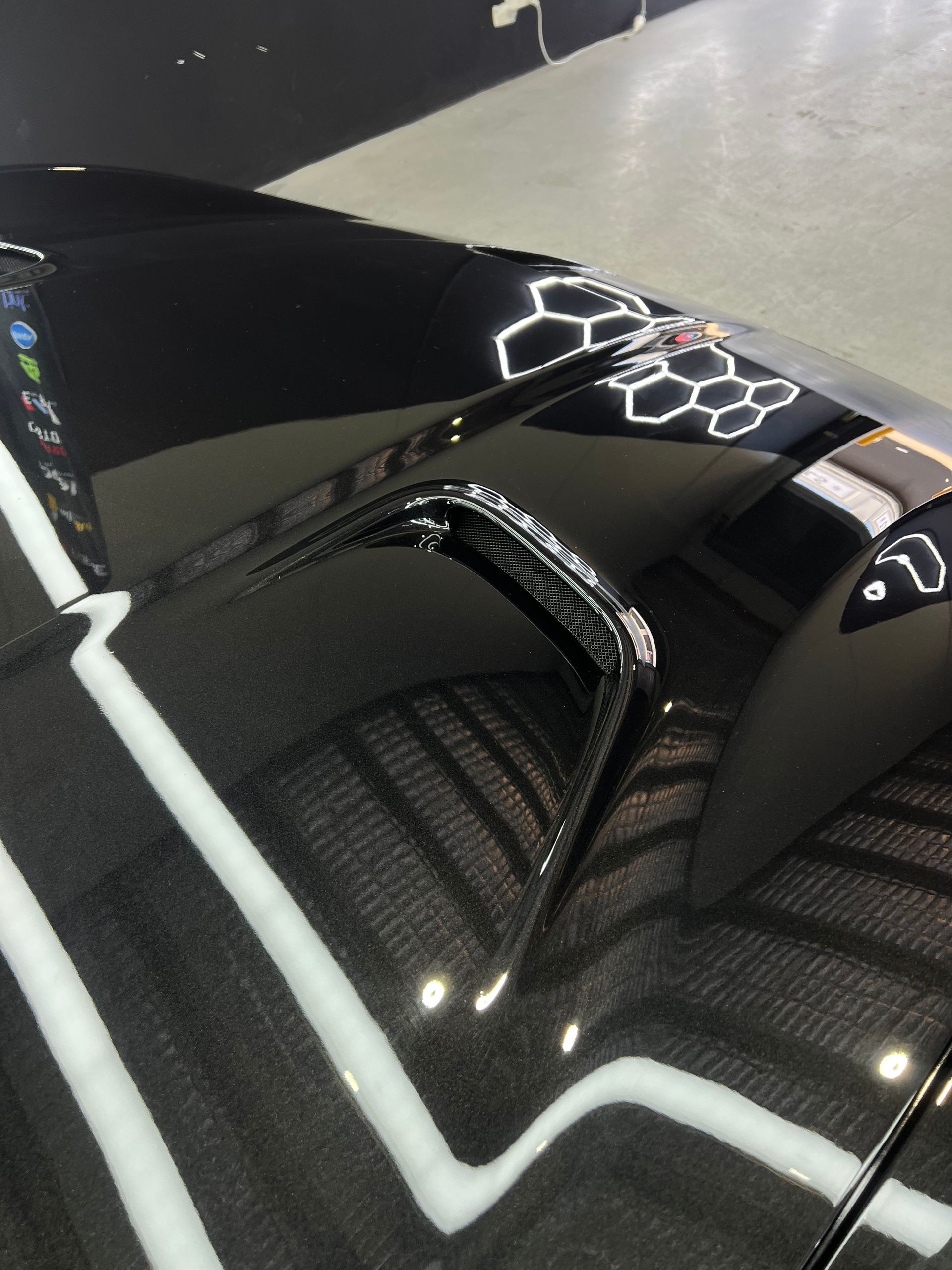 Black car hood with air vent and carbon fiber pattern, reflecting bright white lights.