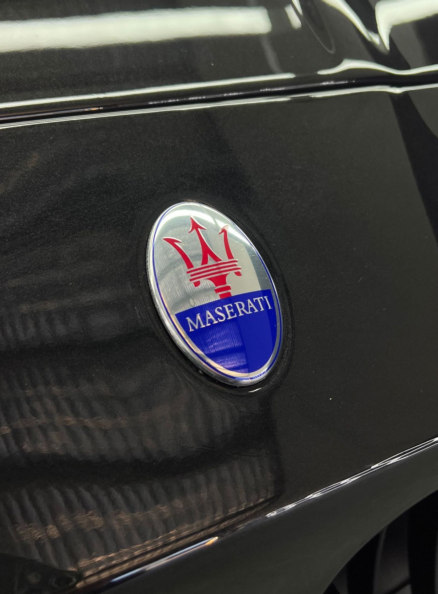 Maserati emblem on a dark grey car; oval shape, red trident, blue and white detail, chrome border.