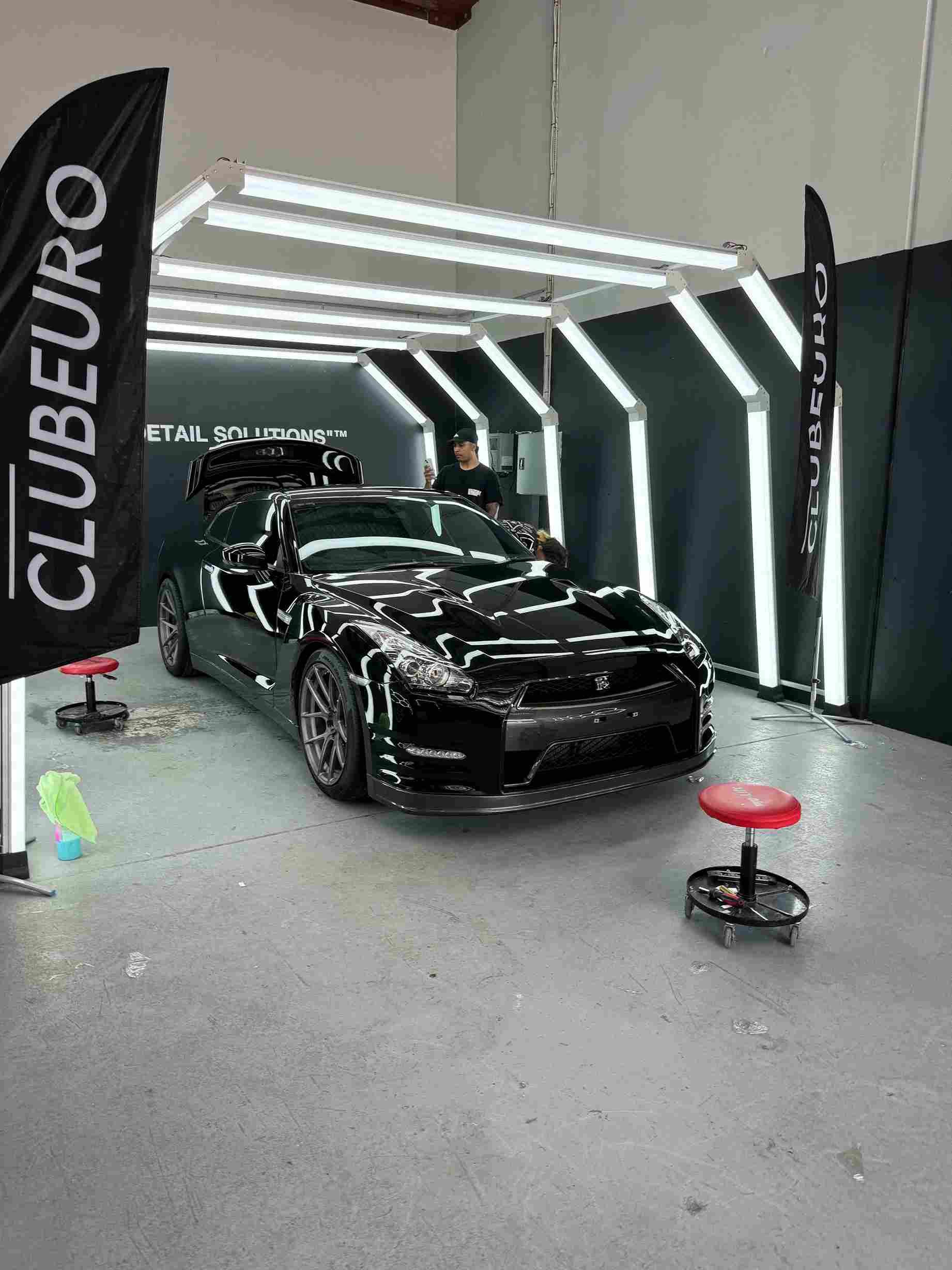 Black sports car under LED lights in a detailing shop, with CLUBEURO flags.