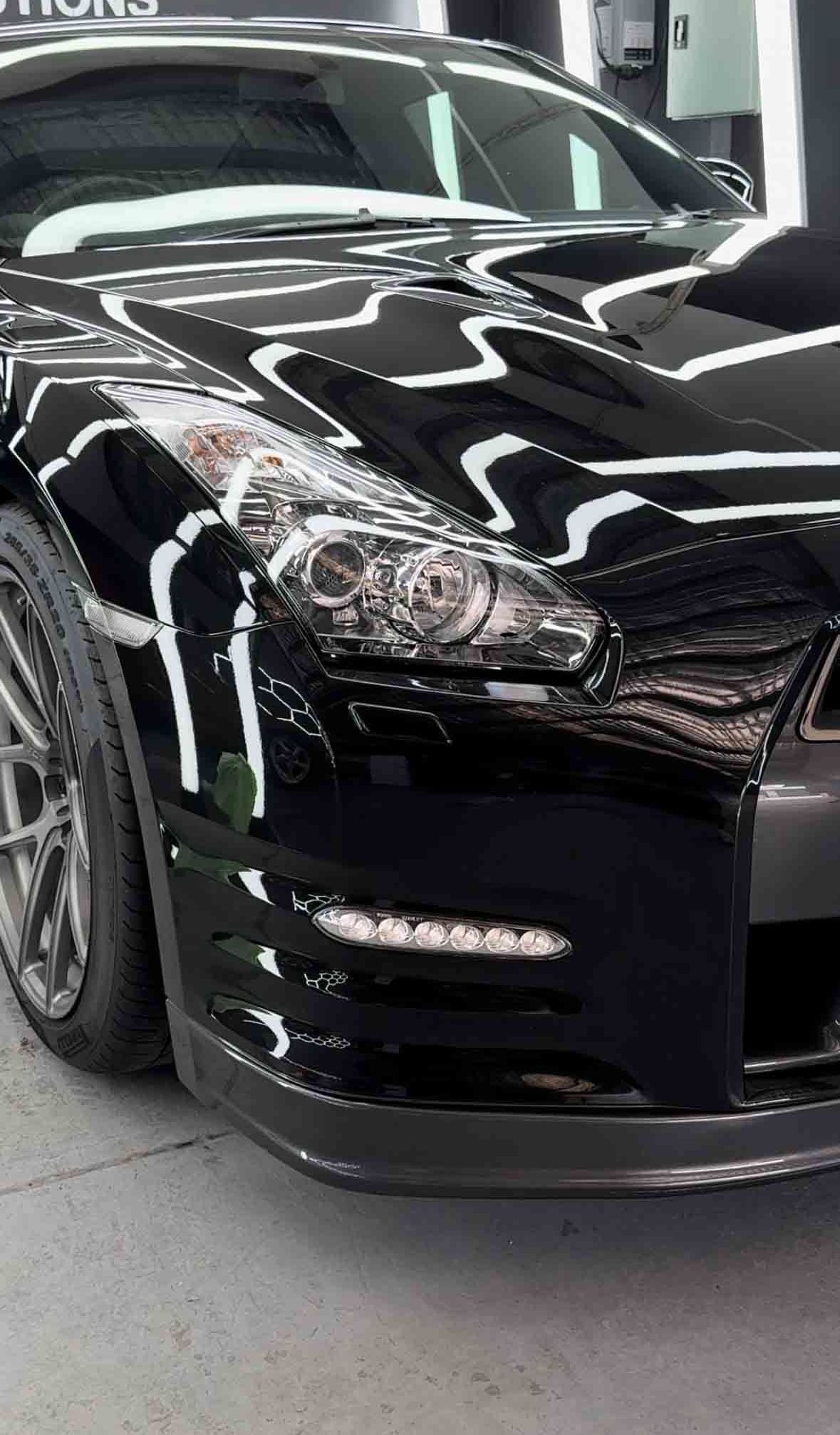 Black Nissan GT-R sports car, close-up of the front. Shiny black paint reflects lights, in a garage.