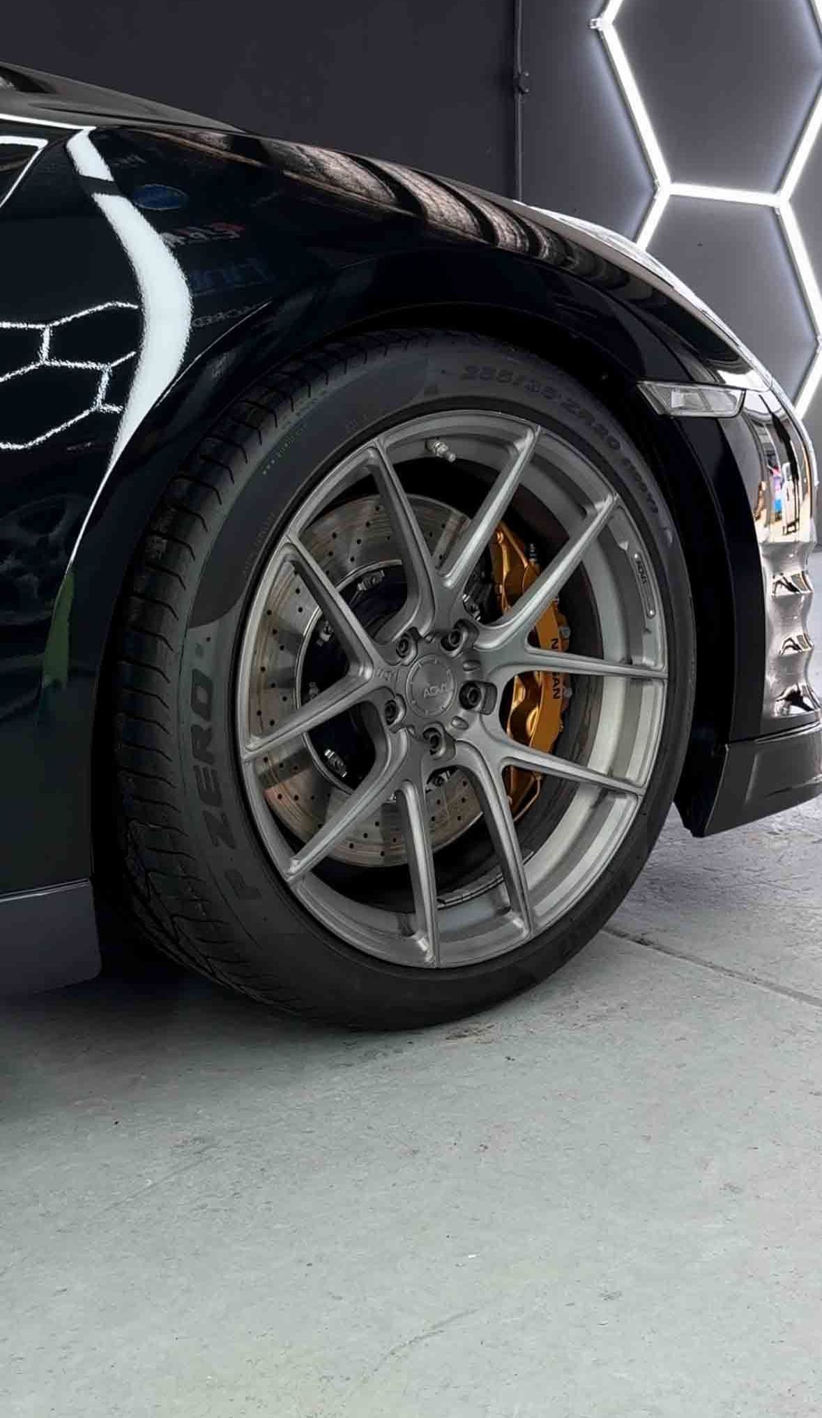 Black sports car with grey rim and orange brake, against a hexagon-patterned wall.