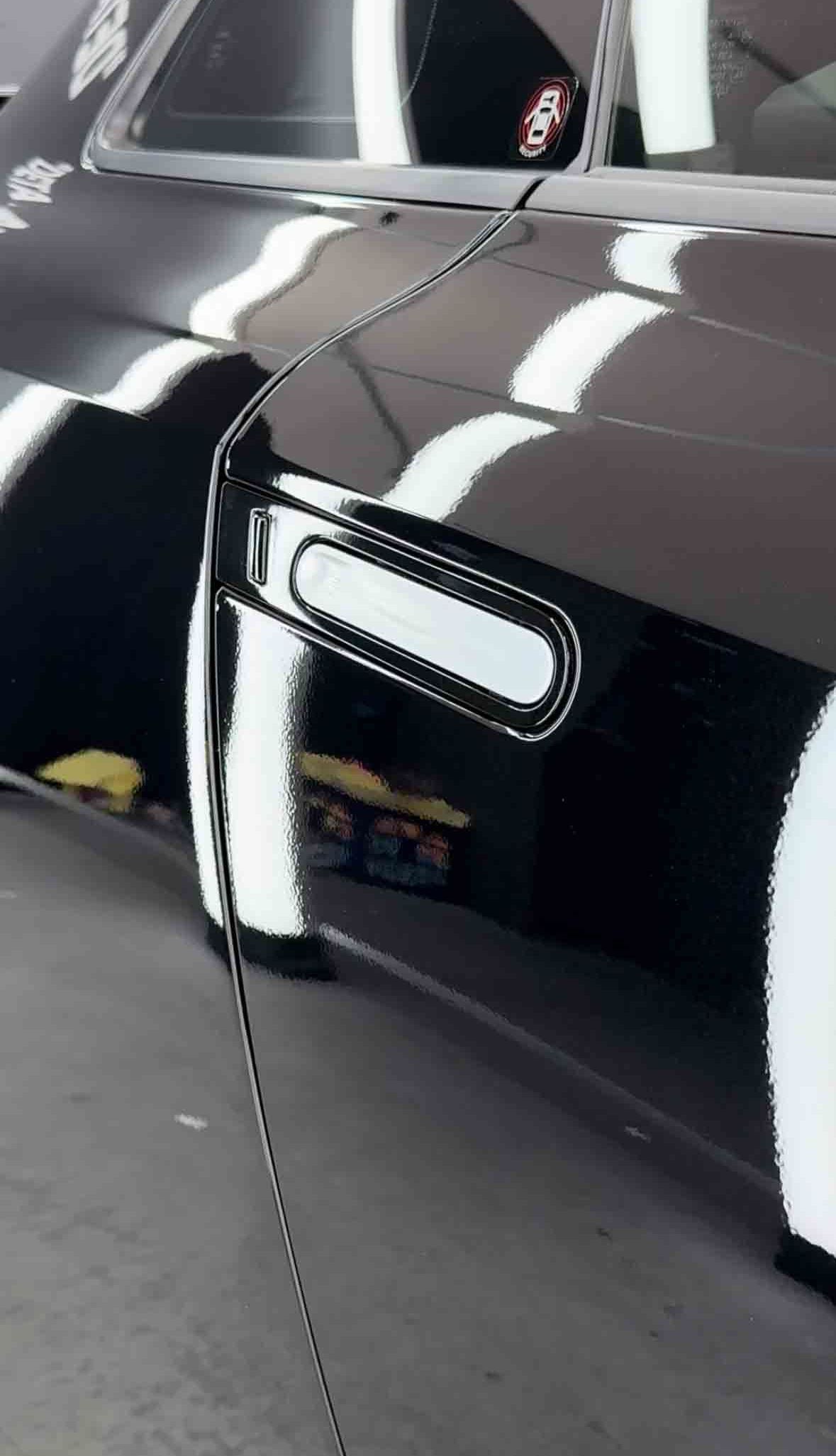 Black car door with chrome trim reflecting a bright overhead light.