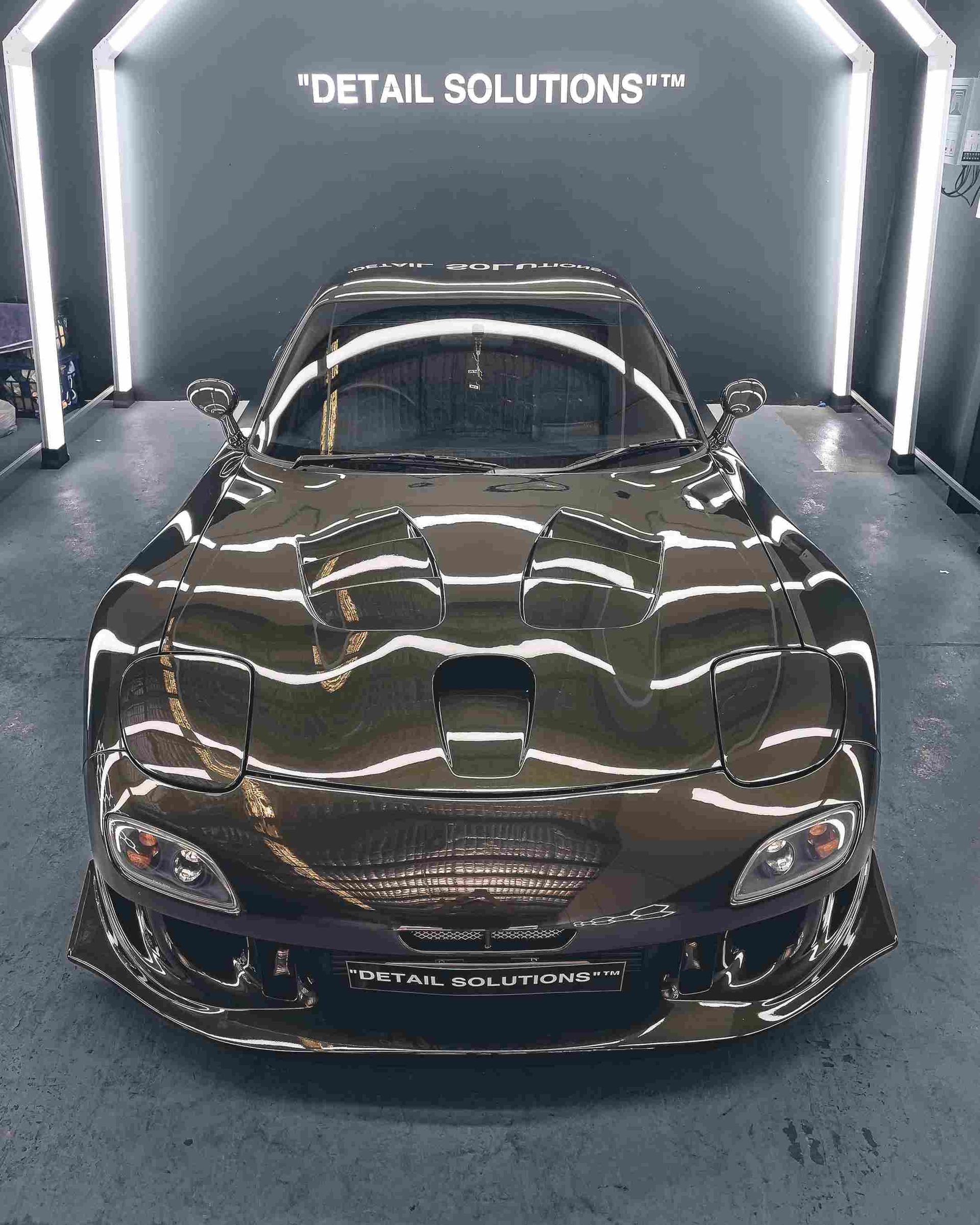 Shiny, gold-colored Mazda RX-7 in a detailing shop, under bright lights.