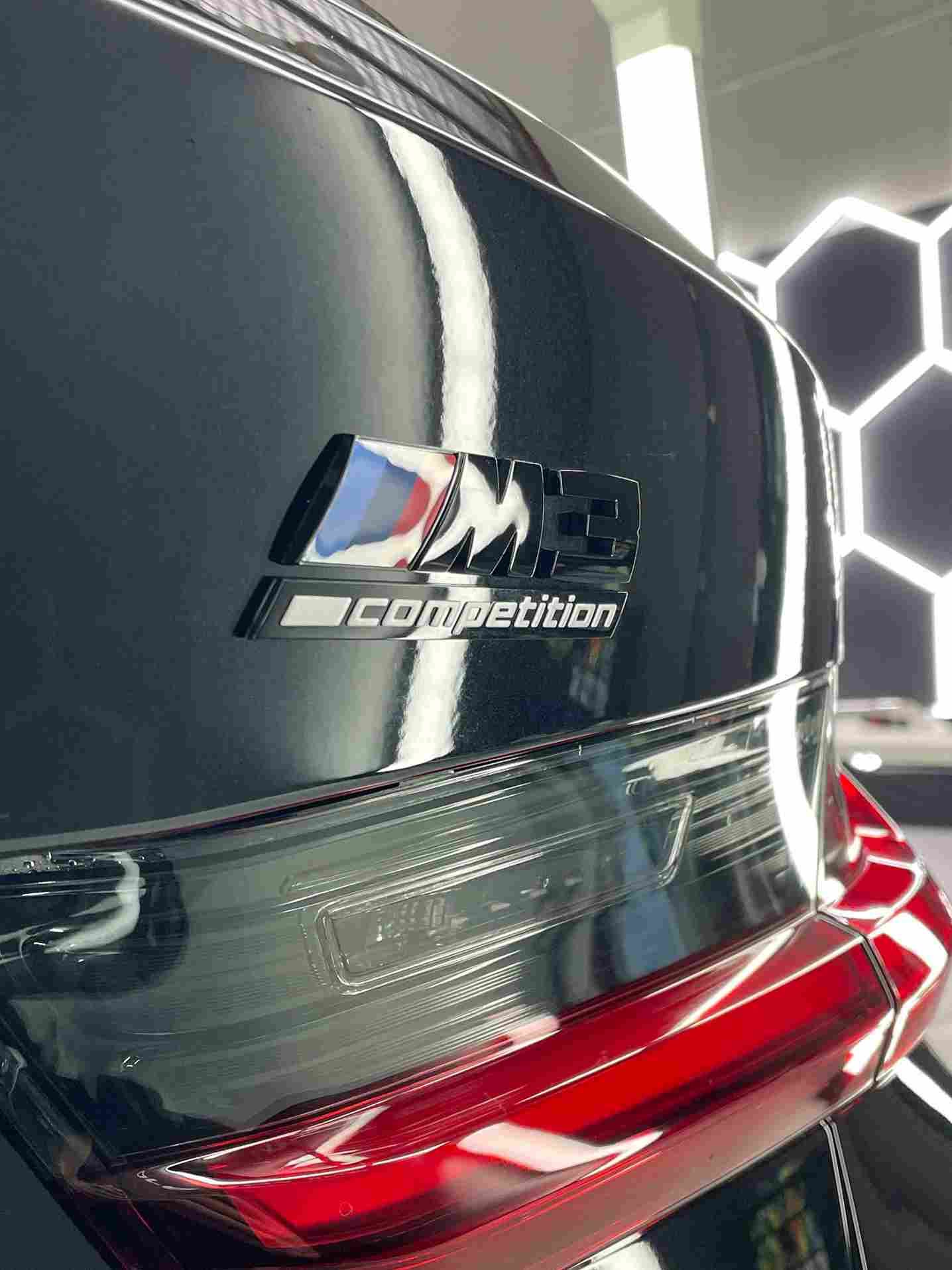 Close-up of a black BMW M8 Competition badge on a car's trunk, with the M logo and red taillight visible.