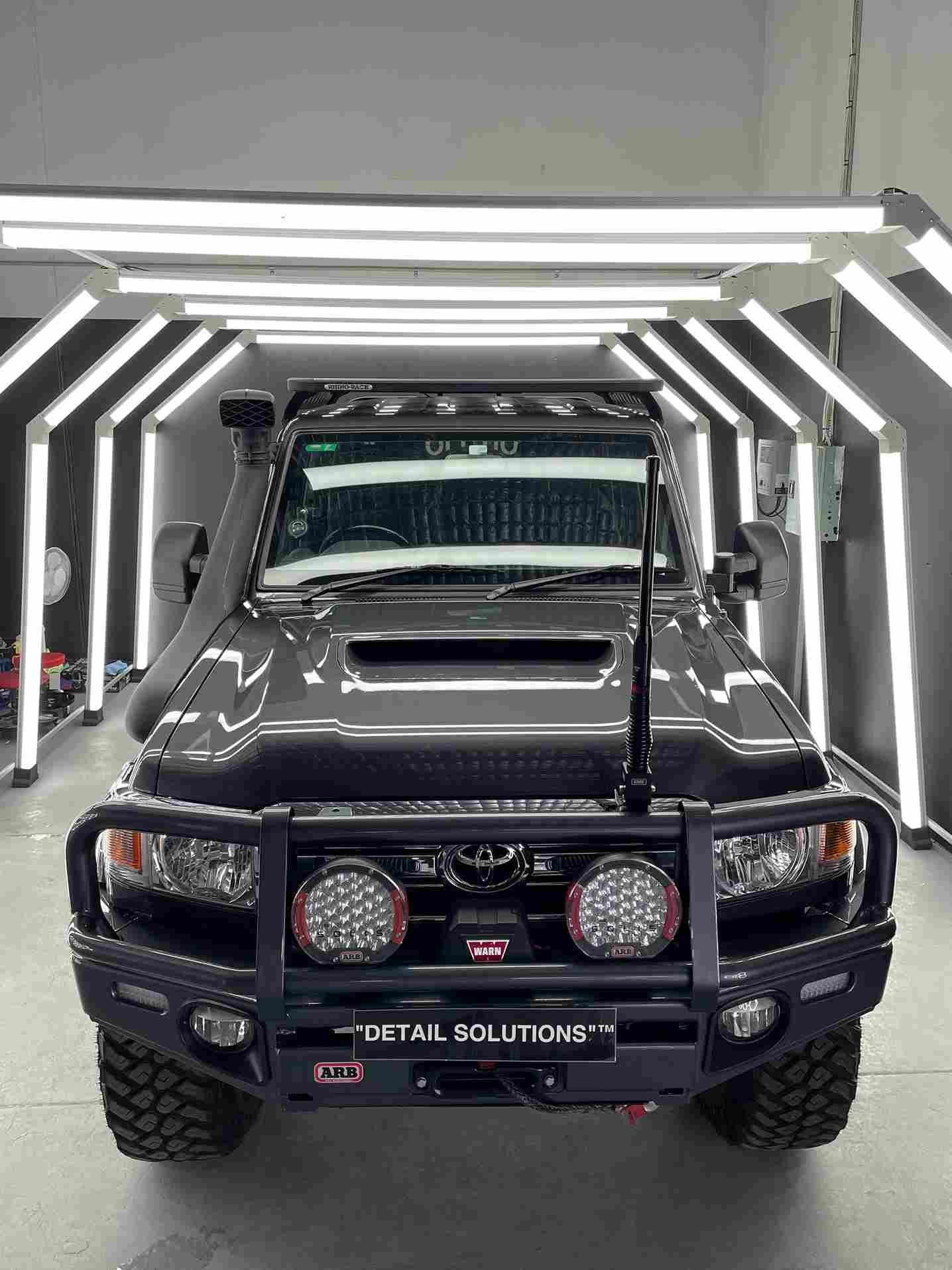 Dark gray Toyota Land Cruiser with off-road modifications, lit by overhead lights inside a detailing shop.