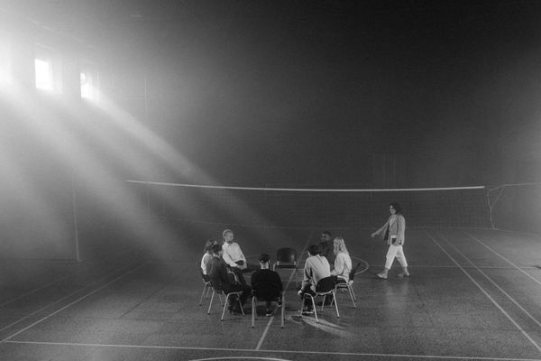Group of people in chairs, one walking. Dimly lit gymnasium with sunlight streaming in.