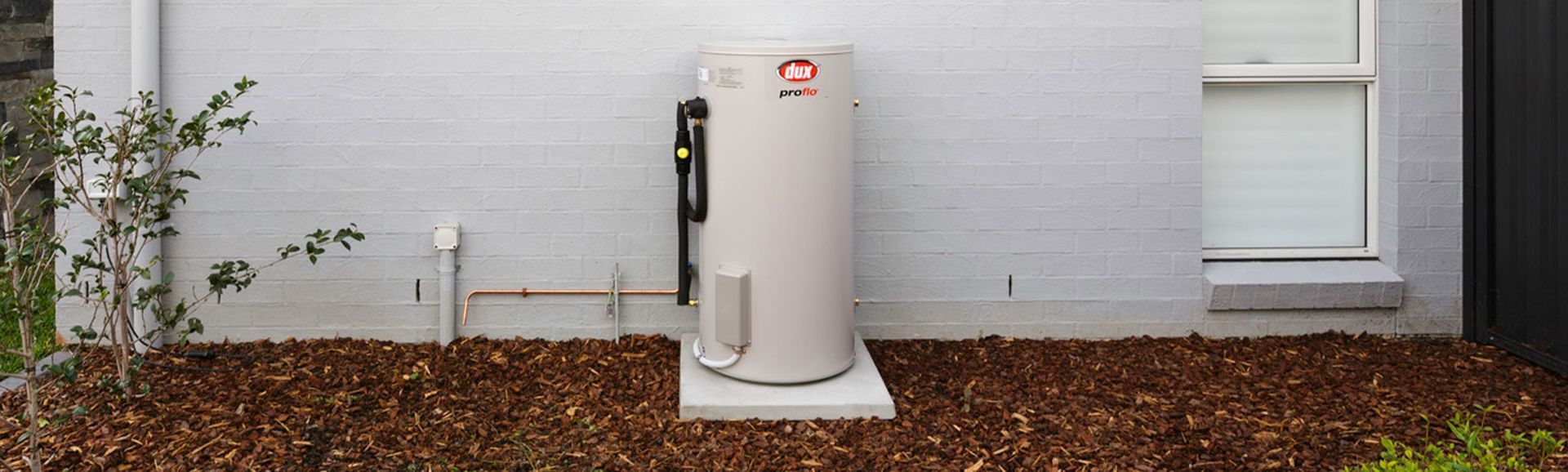 Outdoor Water Heater Unit Beside a Brick Wall, With White Siding and Brown Mulch Bed — Forster Hotwater in Forster, NSW