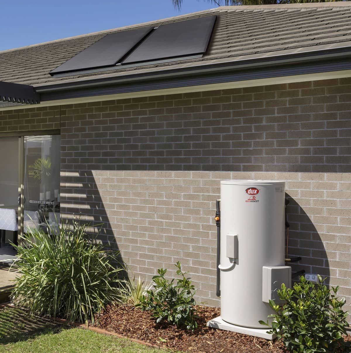 Solar Water Heater Tank Beside a Brick House With Rooftop Panels — Forster Hotwater in Smiths Lake, NSW
