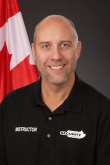 A man wearing a black shirt that says instructor is smiling in front of a canadian flag.