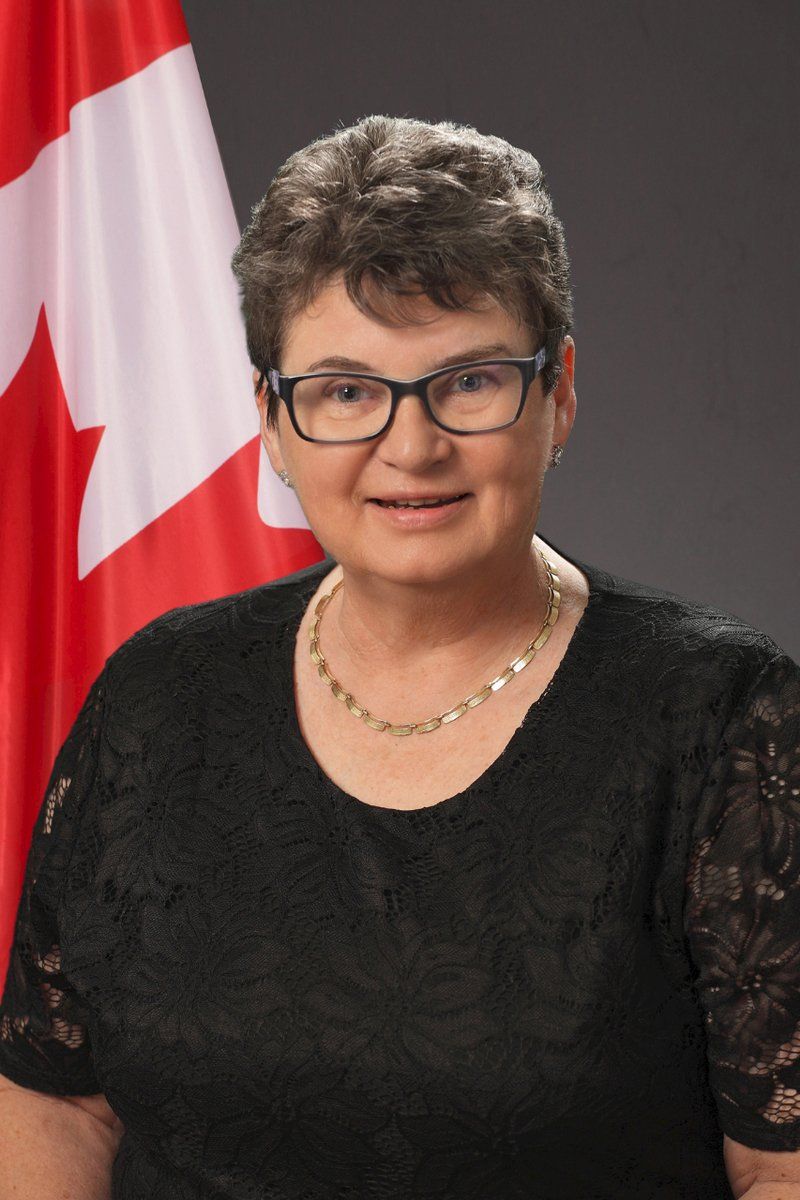 A woman wearing glasses and a necklace is standing in front of a canadian flag.