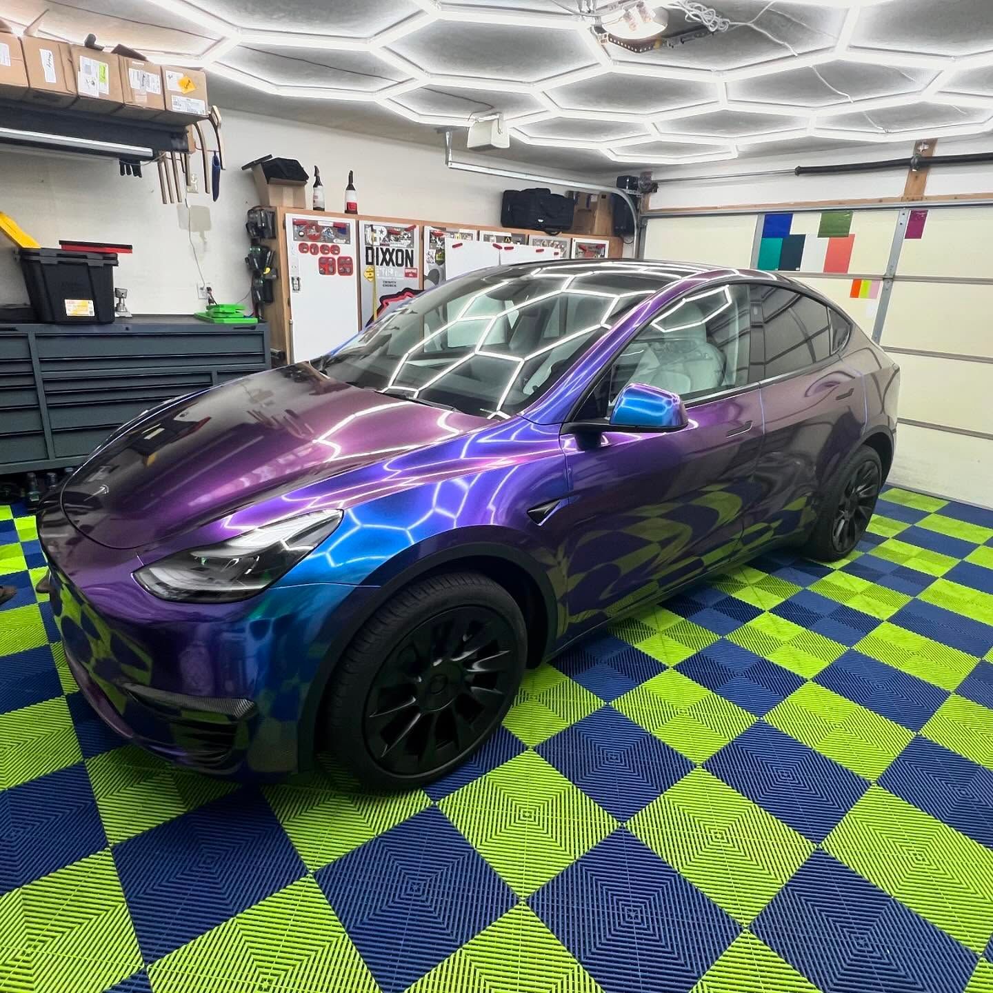 Purple and blue Tesla Model Y in a garage with honeycomb lighting and checkered floor.