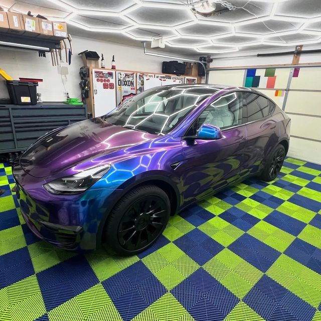 Tesla with a purple and blue iridescent wrap parked in a garage with hexagon lights and a checkered floor.