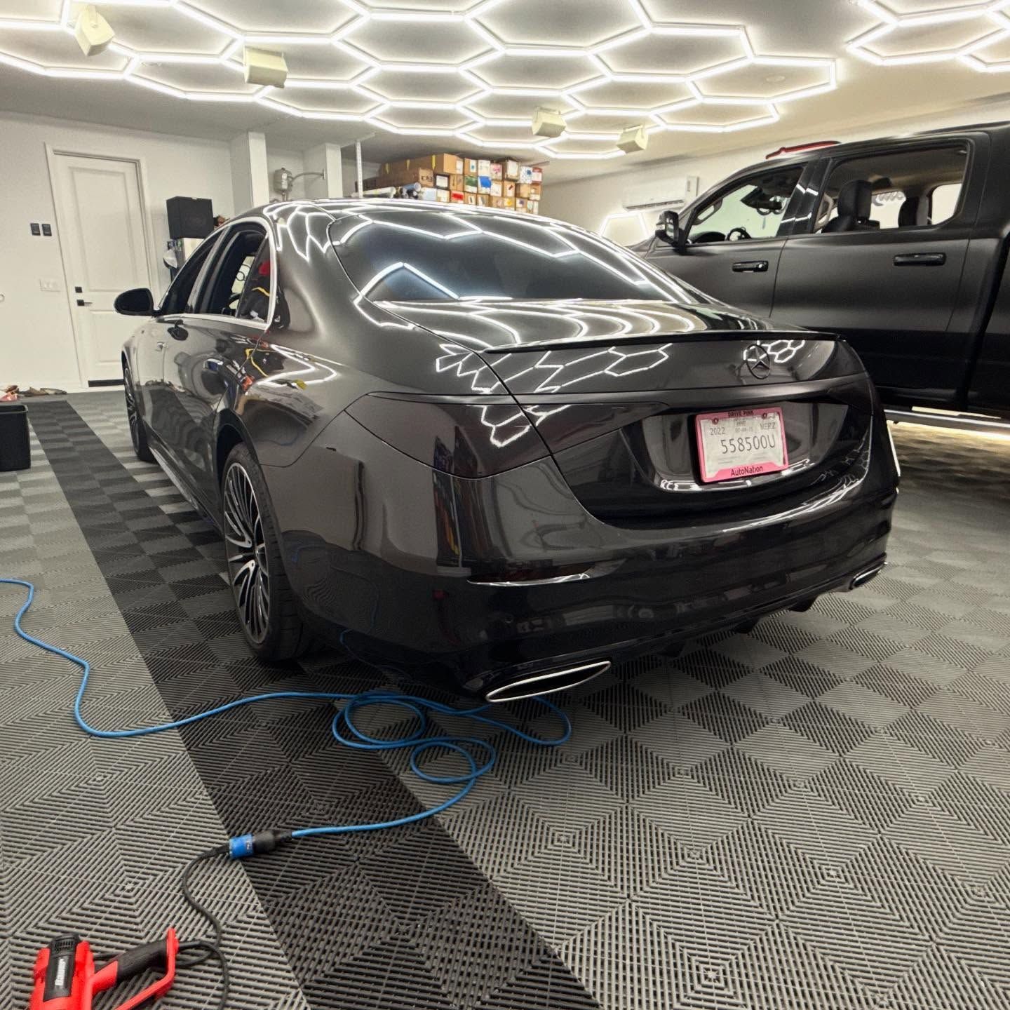 Black sedan in a garage with tiled floor, under hexagon lights, next to a black truck.