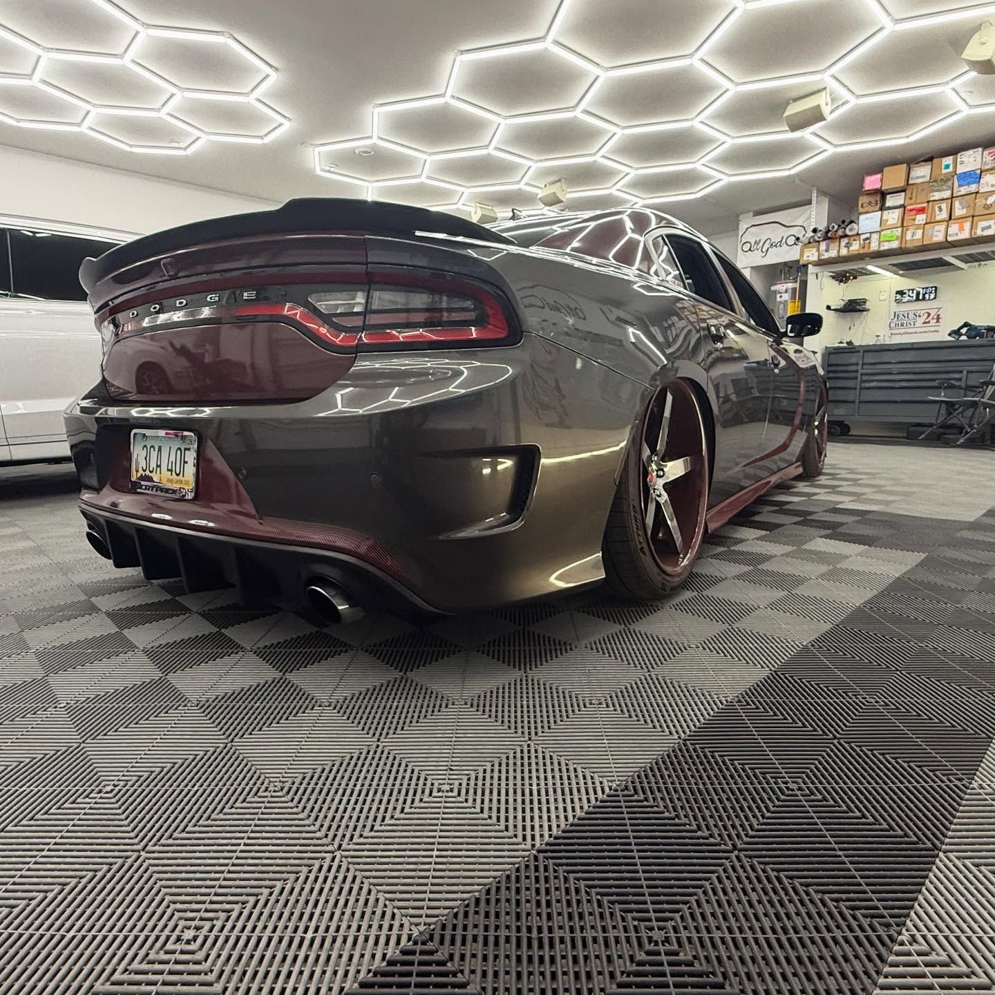 A dark gray Dodge Charger with custom red-tinted wheels and a rear diffuser parked in a garage with hexagon LED lighting.