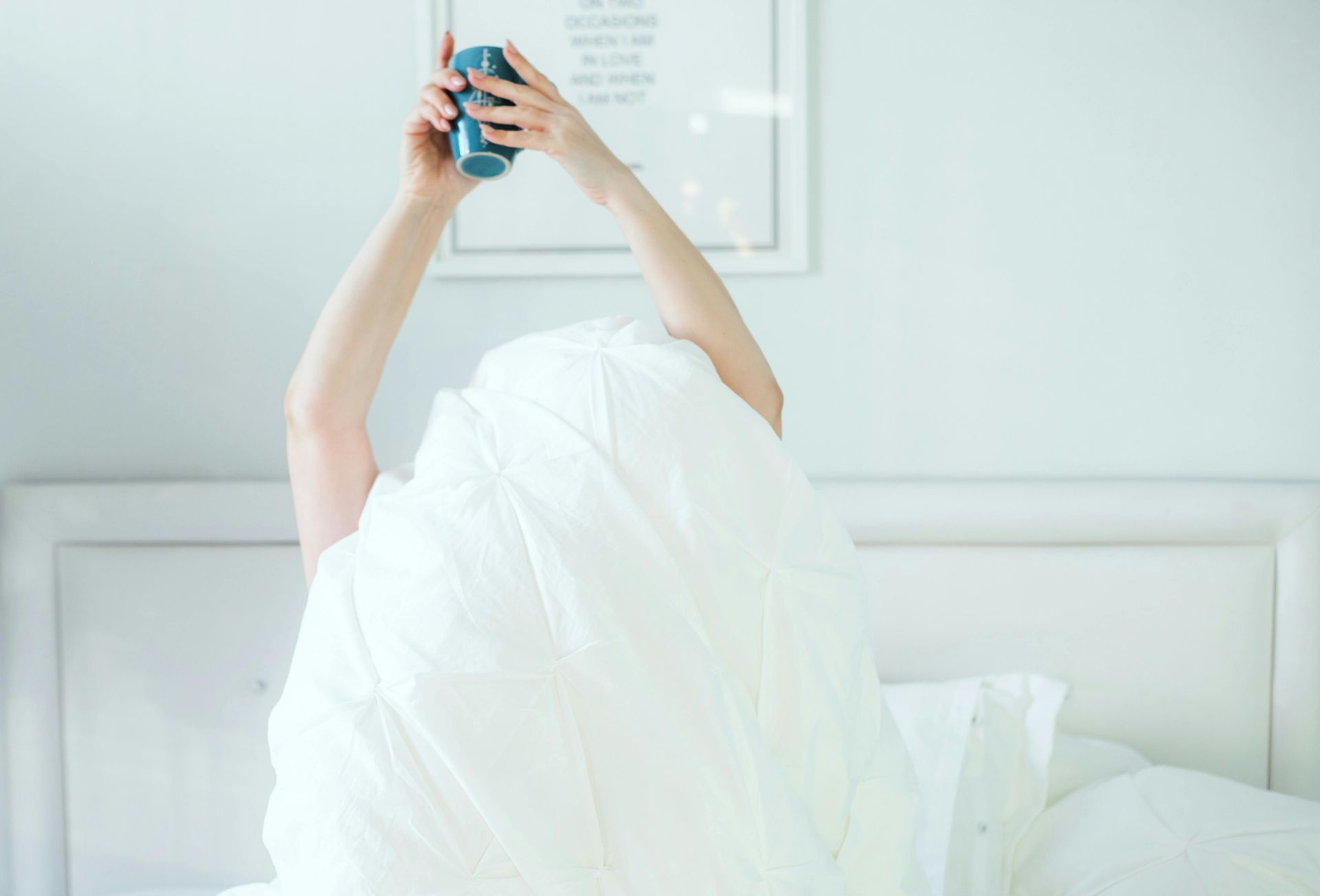 A person sits up in a white bed, covered by a white duvet, holding a blue mug above their head.