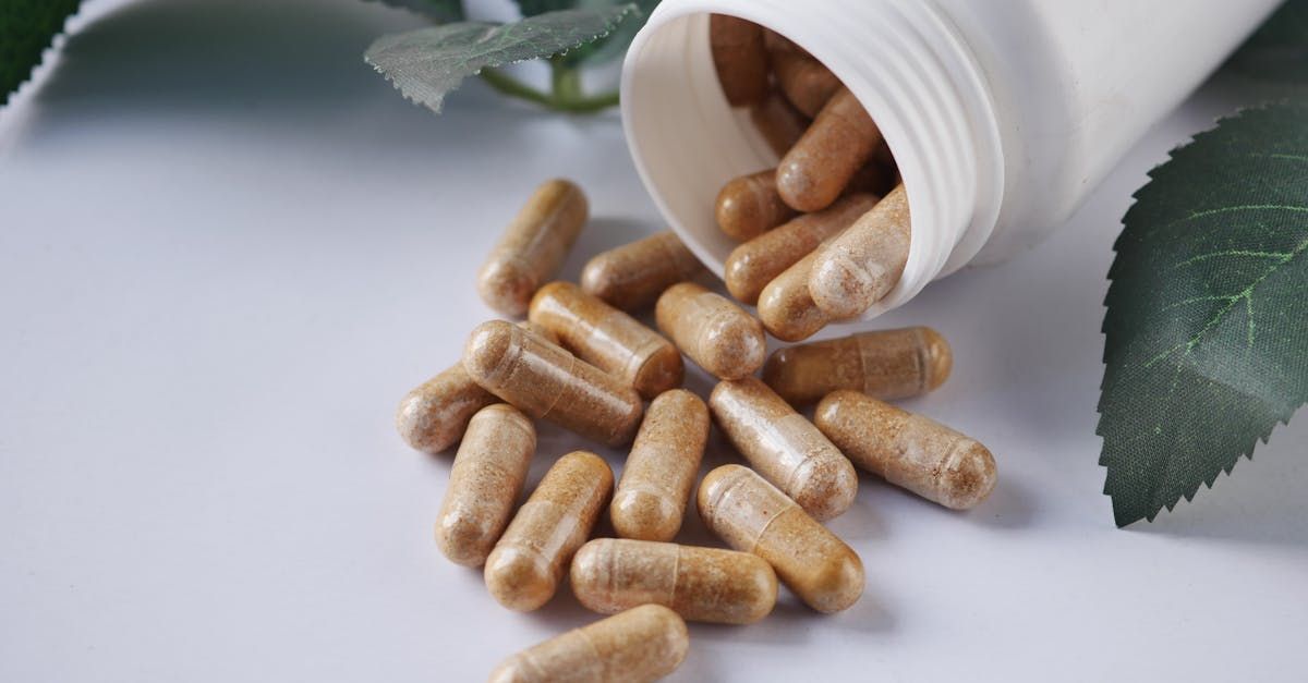 A white bottle spilling tan powder capsules onto a surface next to green leaves.