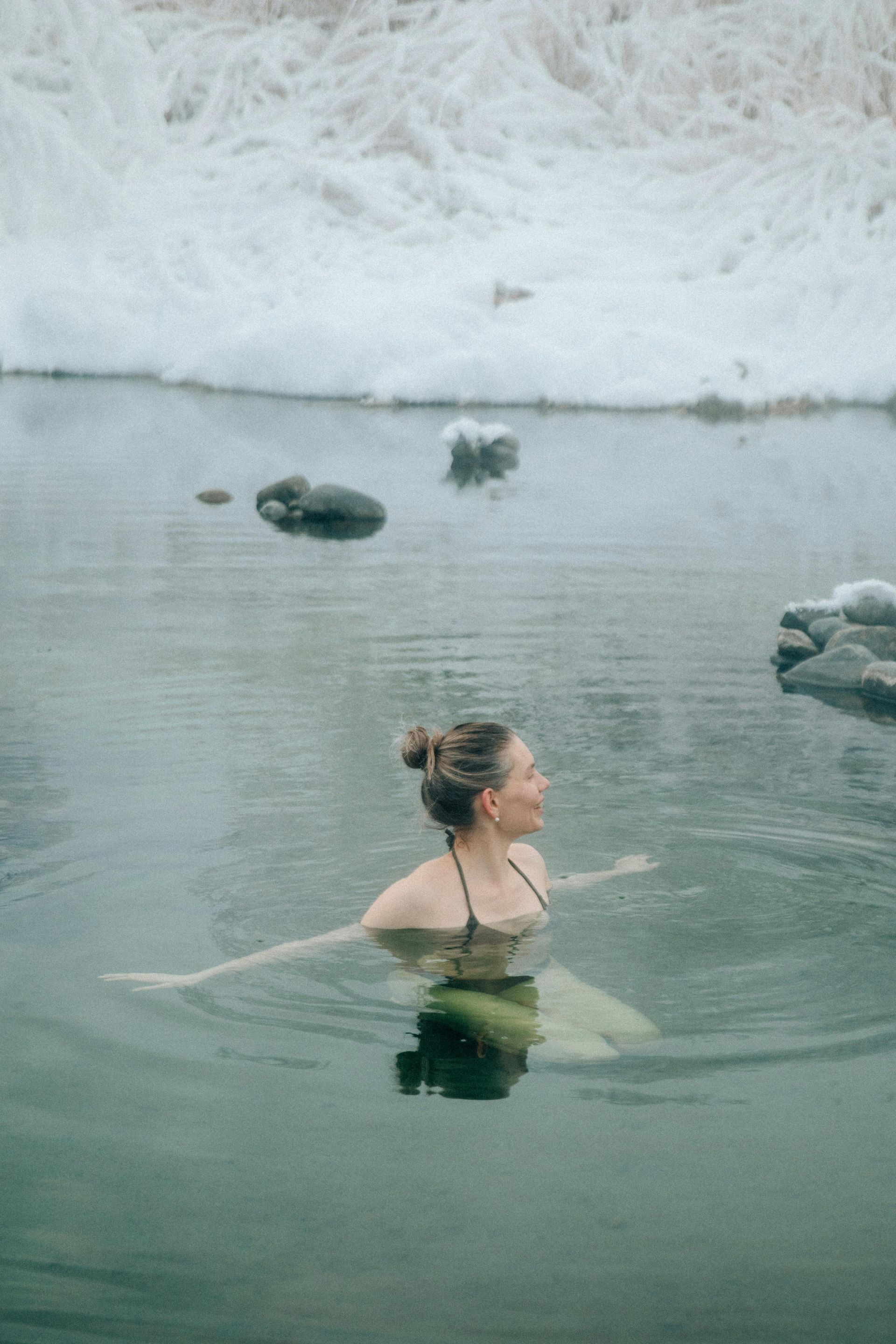 A person with their hair in a bun swimming in a calm, gray geothermal pool surrounded by snow and dark rocks.