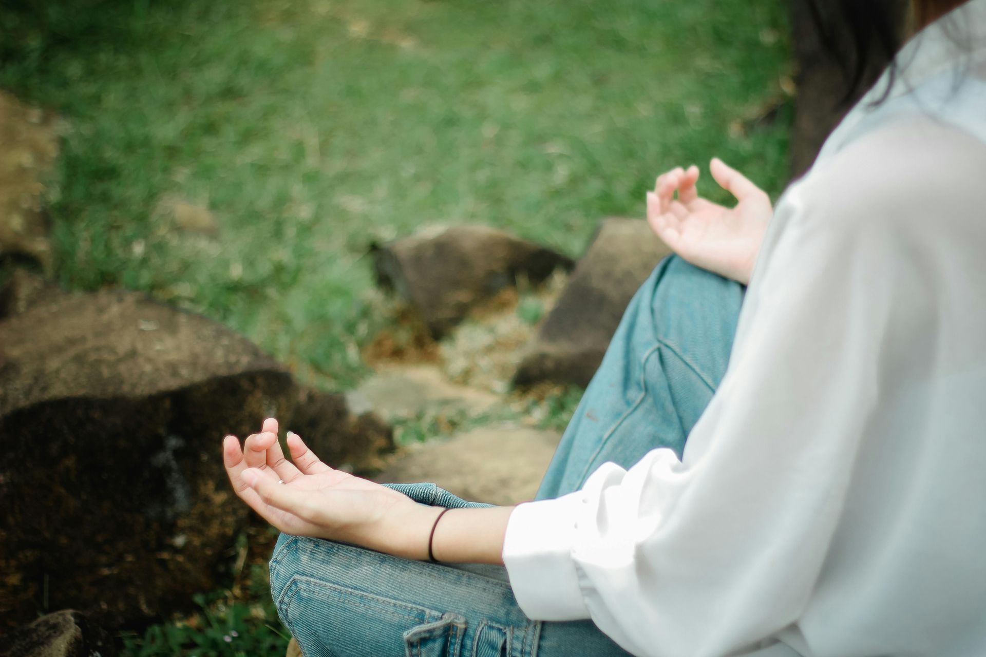A person sitting in a meditation pose, hands open on their knees, outdoors near rocks and grass.