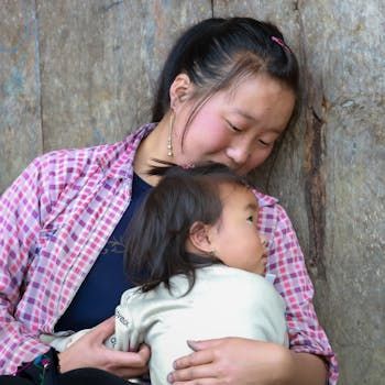 A person in a pink plaid shirt leans against a wall, gently embracing a young child resting against their chest.