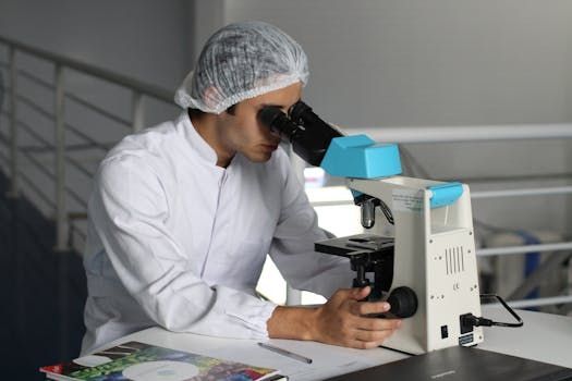 A person in a white lab coat and hairnet looking through a microscope at a desk in a laboratory setting.