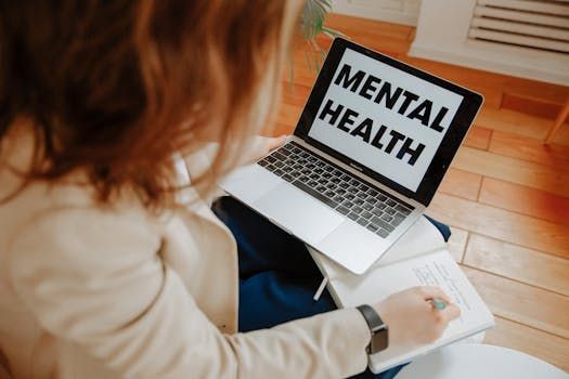 A person sits on a wooden floor, writing in a notebook while looking at a laptop screen that says MENTAL HEALTH.