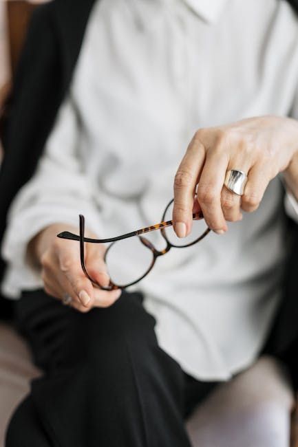 A person wearing a white shirt and black jacket holds tortoiseshell glasses, with a prominent silver ring on their finger.