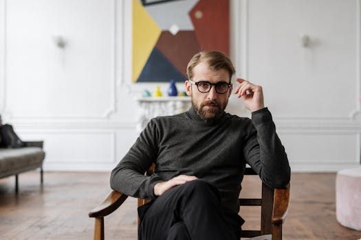 A person with a beard and glasses sits in a wooden chair, hand raised to their temple, in a room with abstract art.