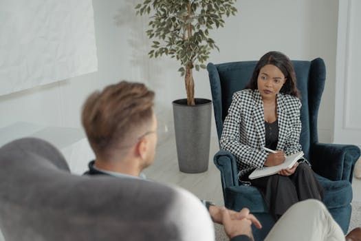 A person in a professional office setting takes notes while sitting in a chair, listening to someone facing away.