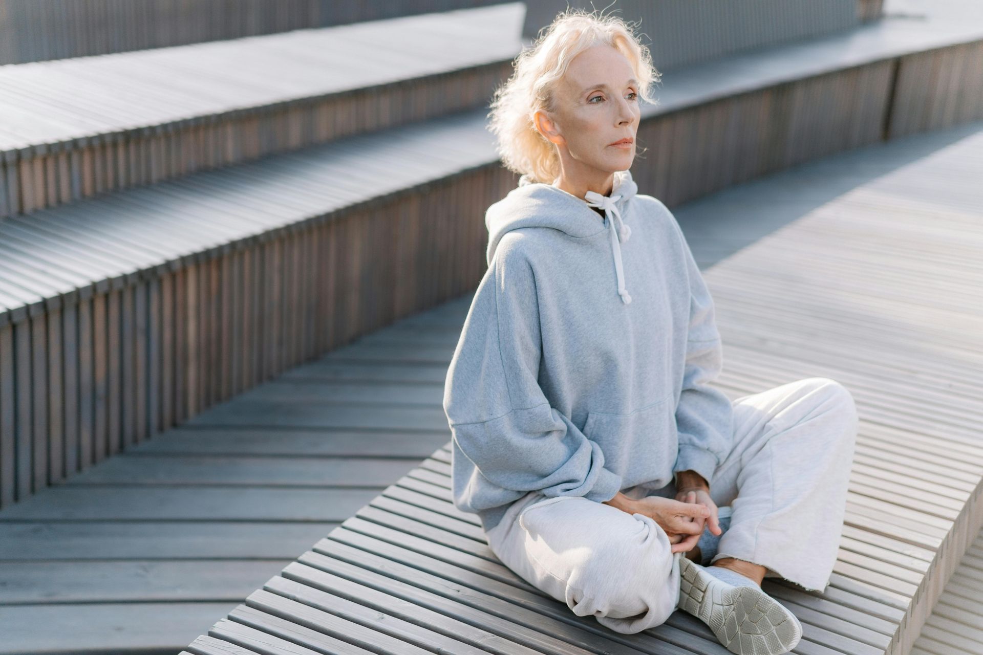 A person sitting in a meditative pose on wooden steps, wearing a light gray hoodie and matching pants.